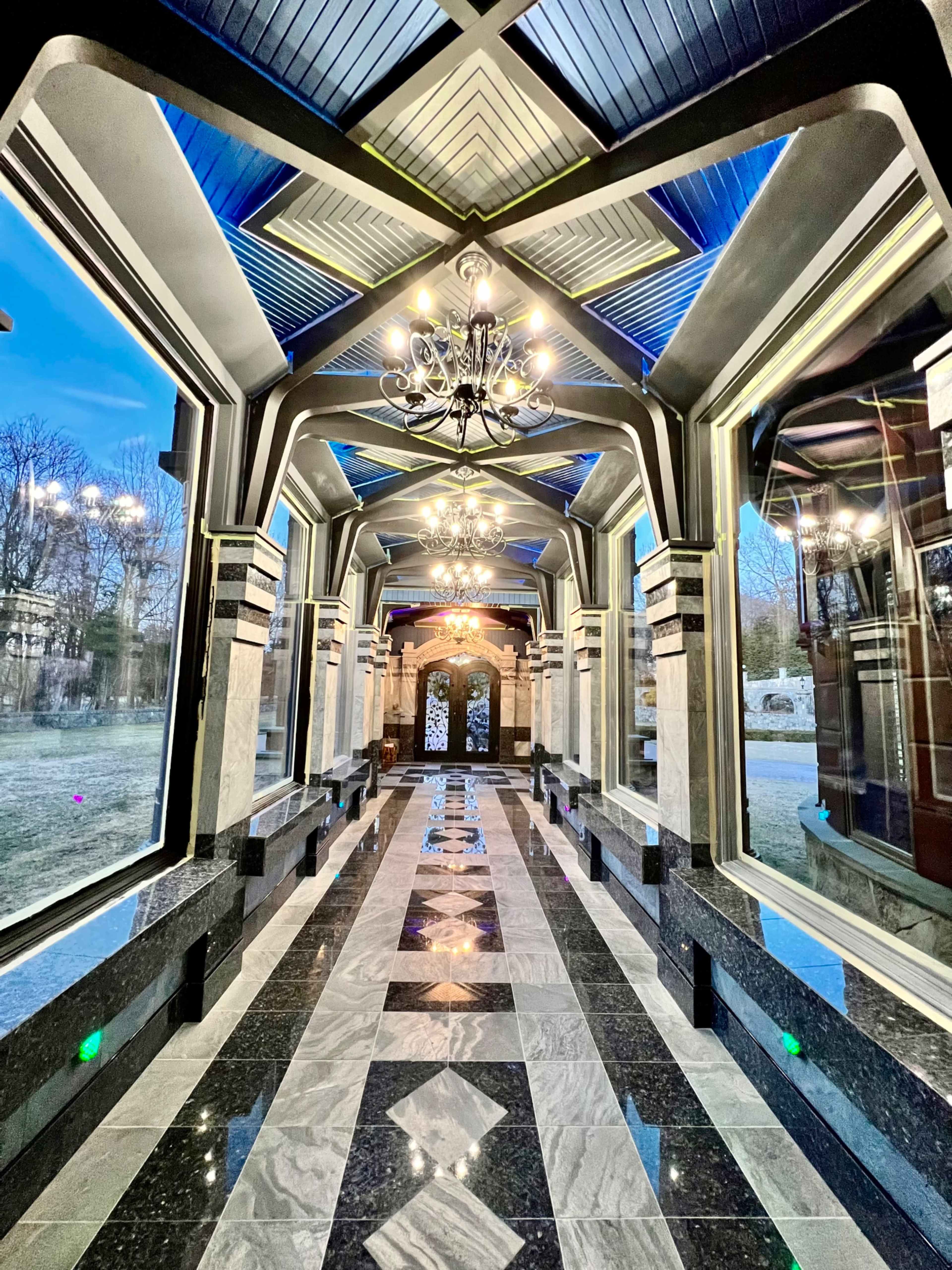 A long, elegant hallway with marble flooring, decorative chandeliers, and large windows on either side.
