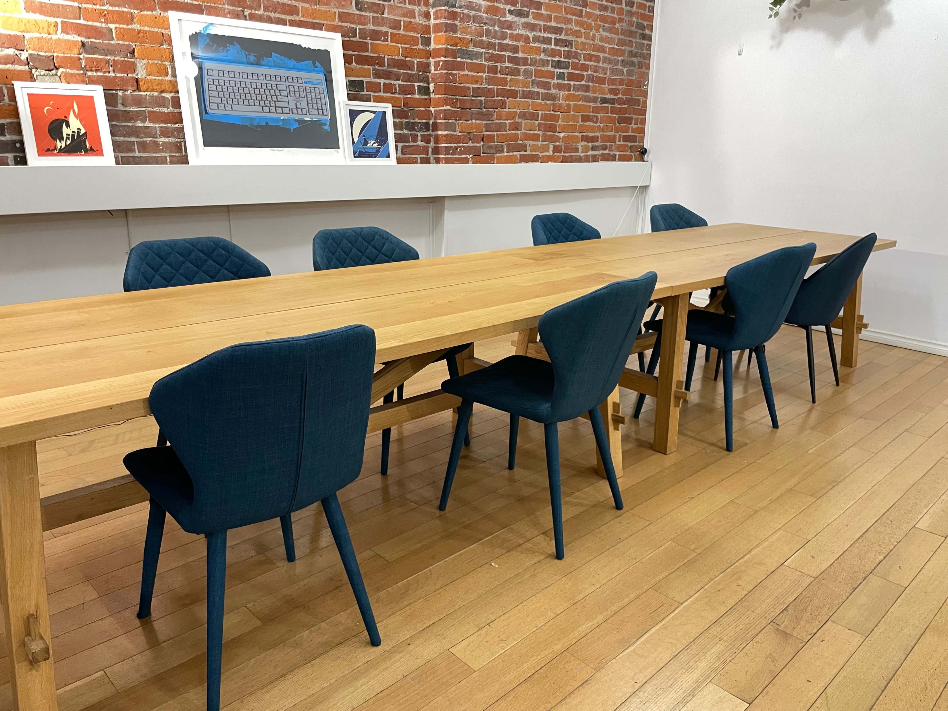 A long wooden table with eight blue upholstered chairs is situated in a room with a brick wall and framed artwork above the table.