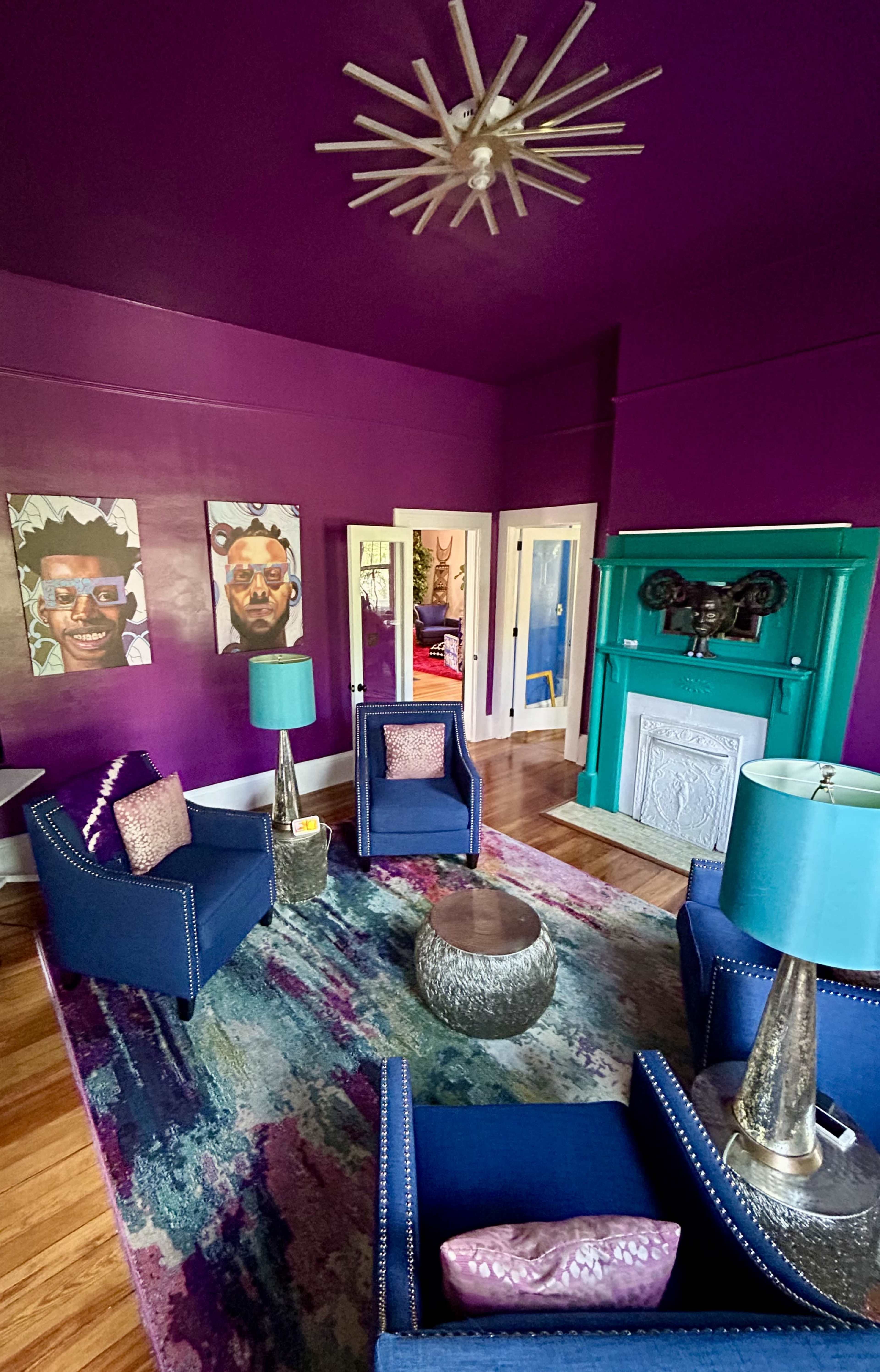The room features a vibrant purple wall, two artwork pieces, a teal fireplace, and modern seating arrangements with decorative lamps.