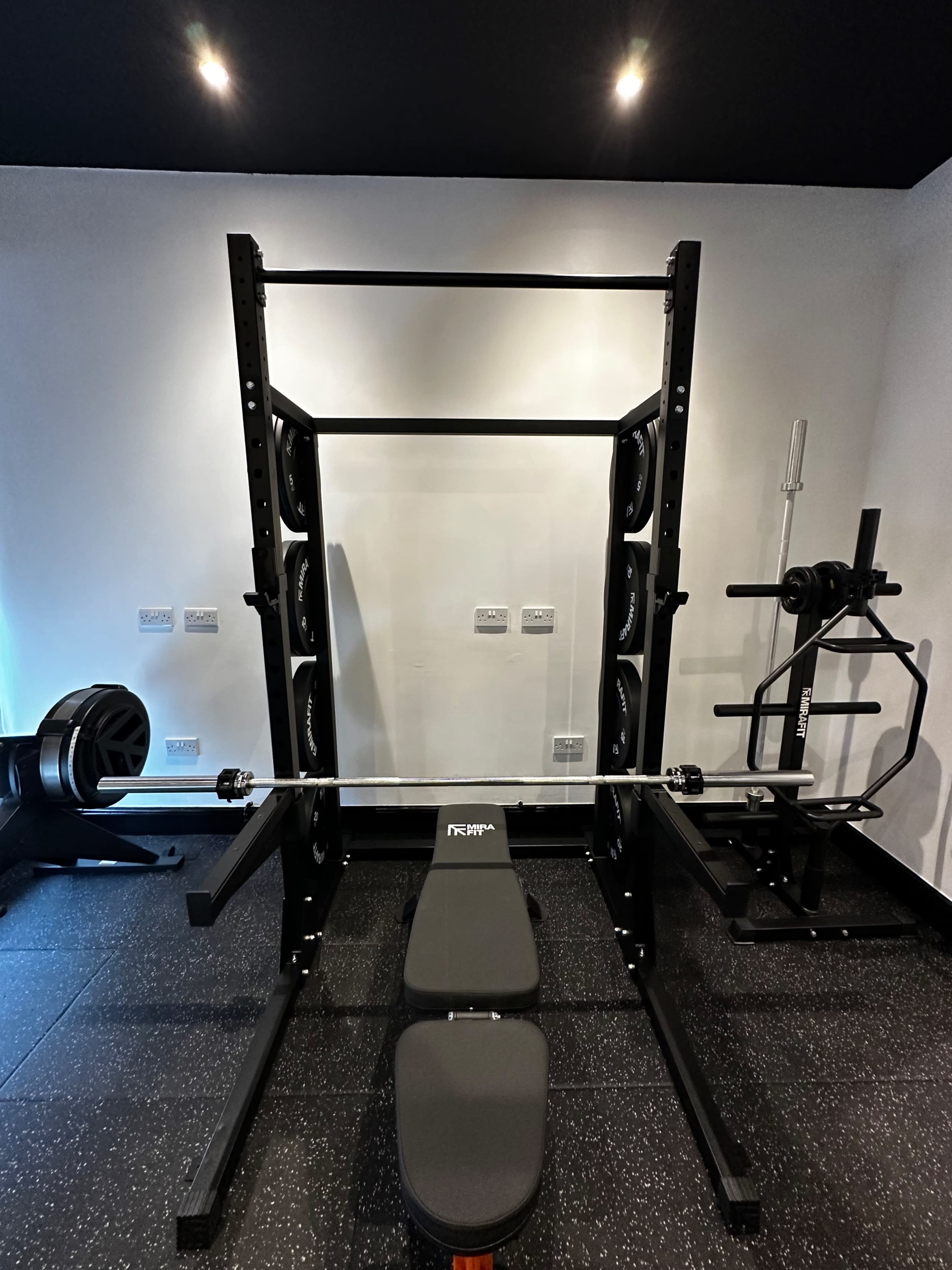 A gym setup featuring a squat rack with weights, a bench, and a barbell alongside additional gym equipment in a well-lit room.