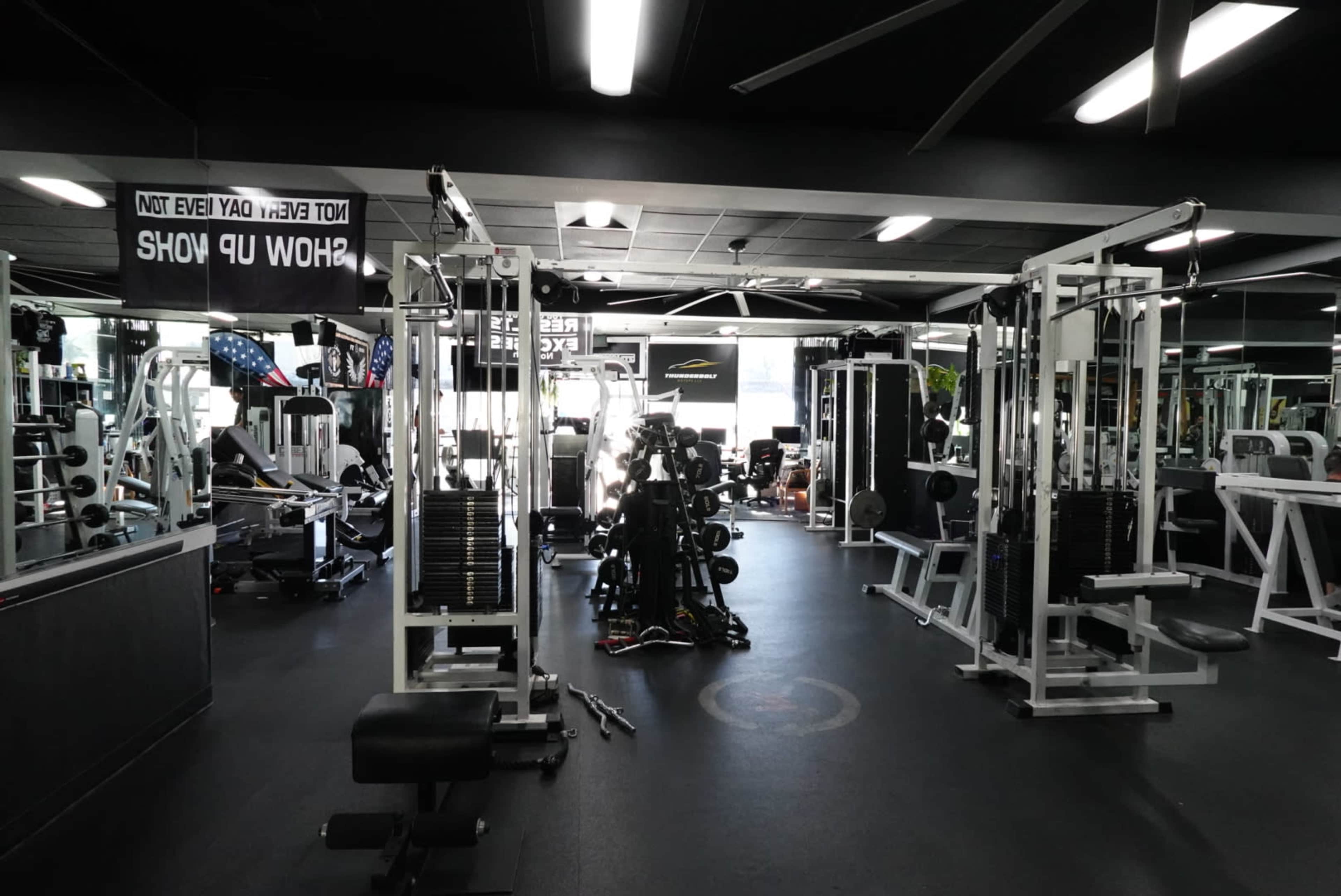 The image shows a fitness gym filled with various weight machines and exercise equipment, with signage hanging from the ceiling.