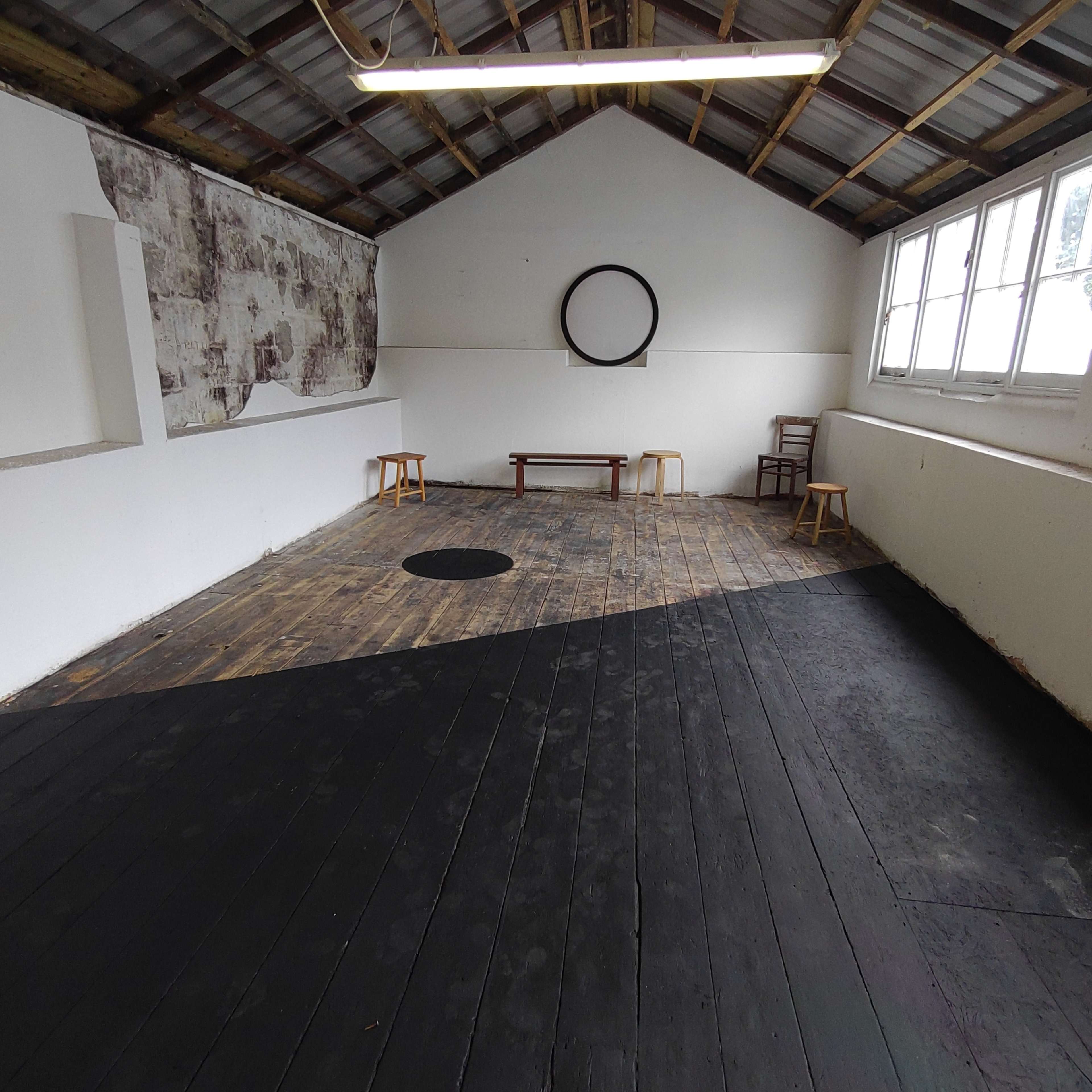 A spacious, partially empty room with wooden floors, a round black mat on the floor, a circular wall decoration, and simple wooden furniture along the walls.