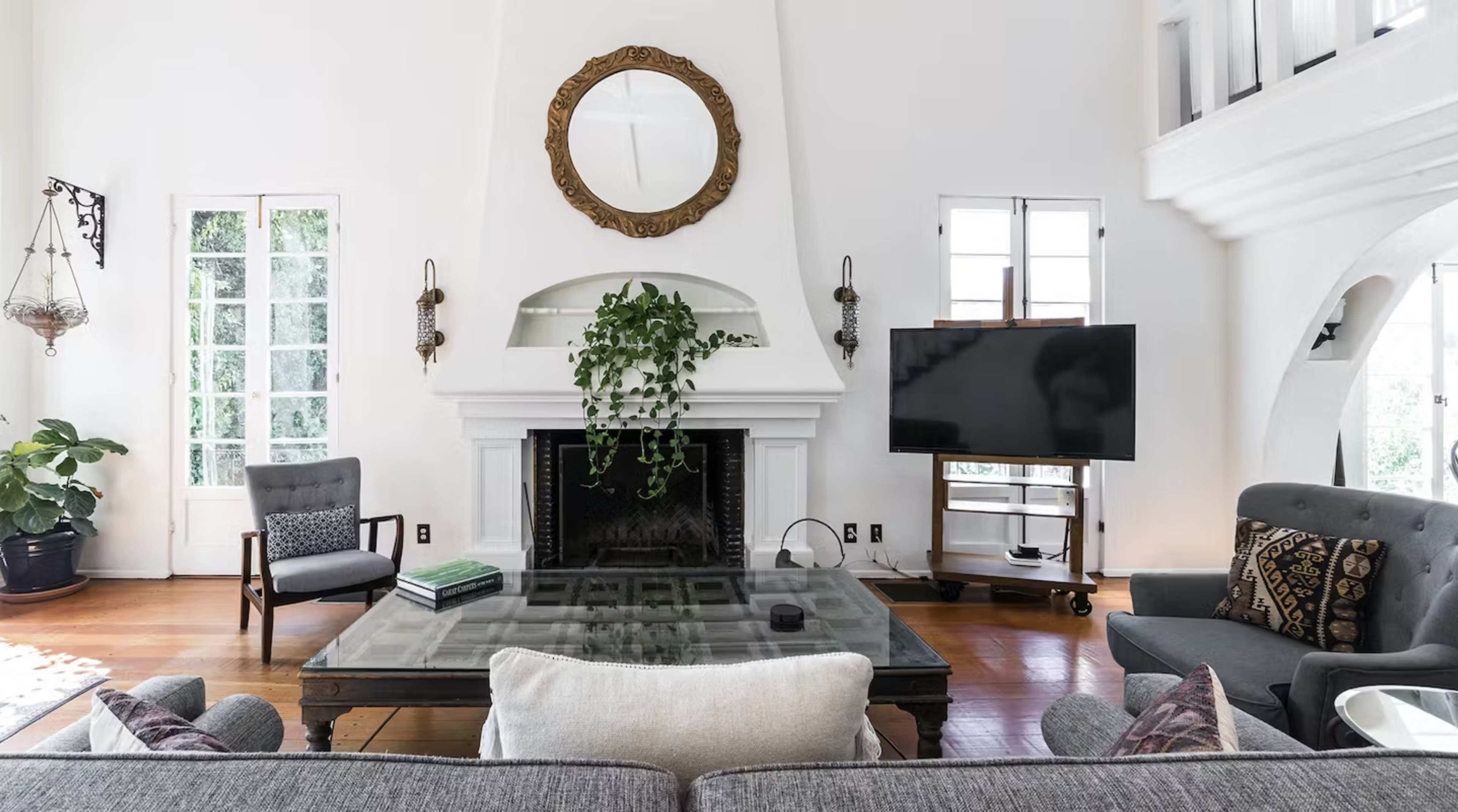 A spacious living room features a fireplace with a large mirror above it, a glass coffee table, and a television beside a potted plant.
