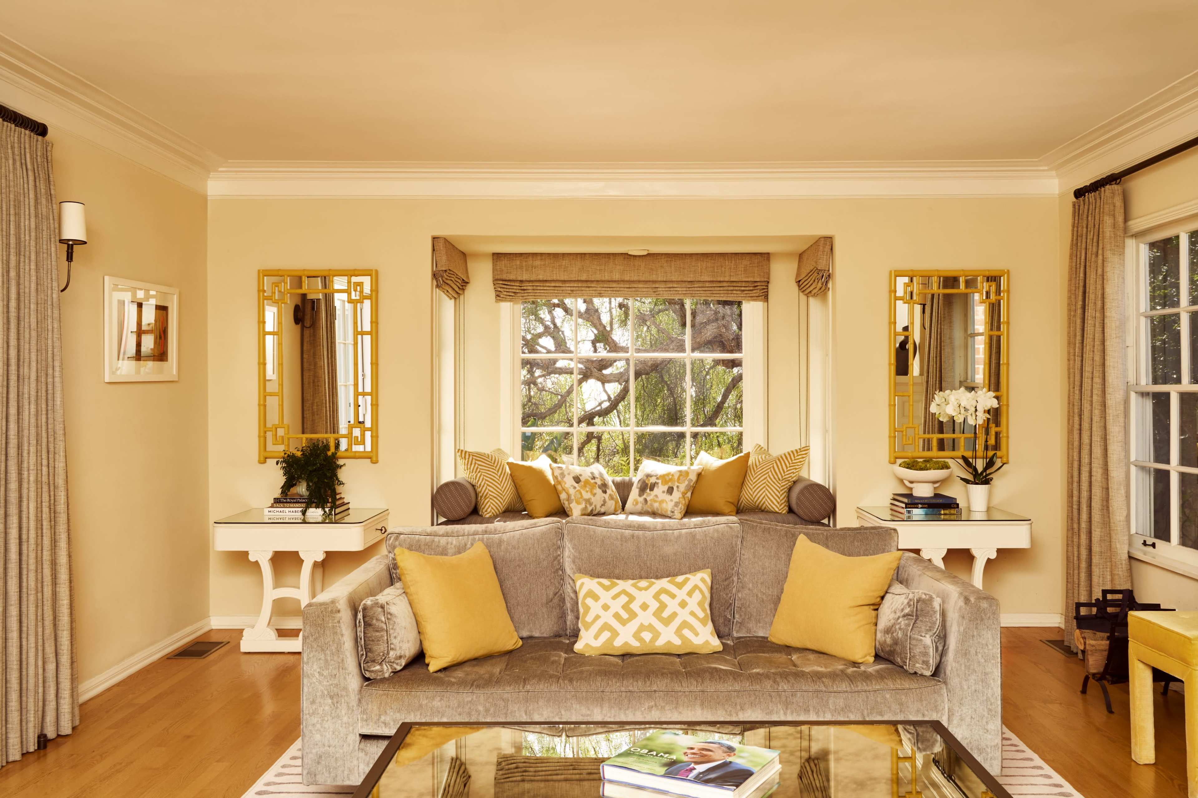 A bright living room features a gray sofa with yellow cushions, two mirrors on the walls, and a window seat with decorative pillows, complemented by wooden side tables.