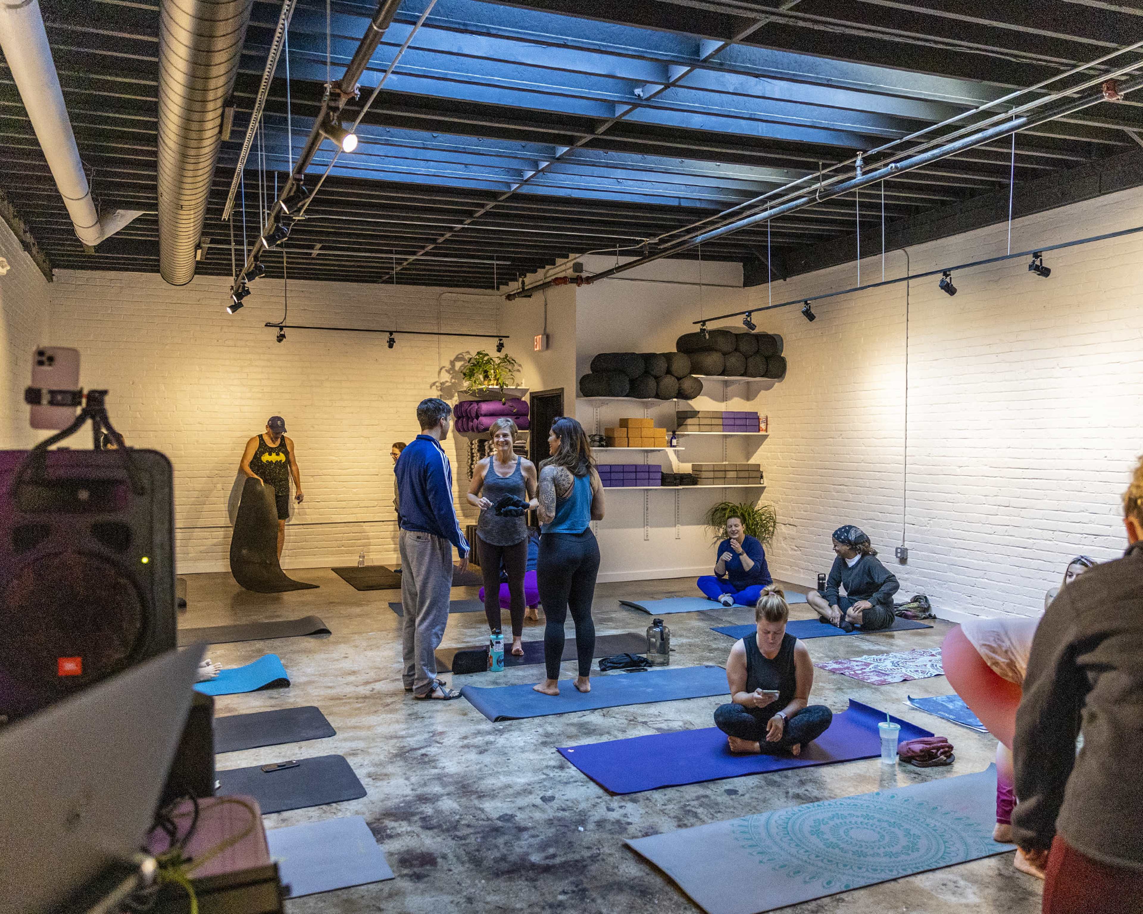 The photo shows a yoga studio with several people practicing on mats while others are engaged in conversation.
