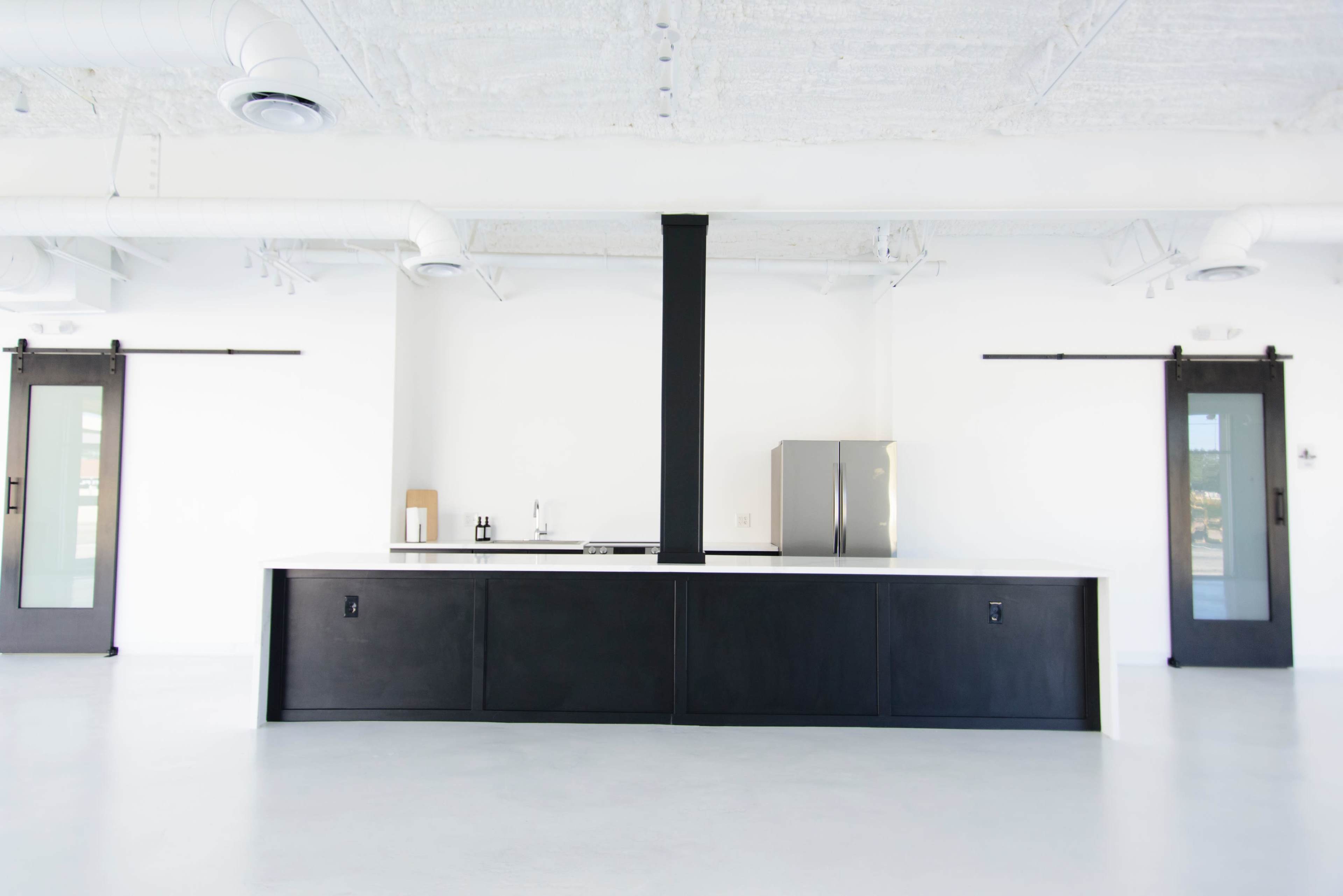 A modern kitchen with a large black island, stainless steel refrigerator, and sliding doors on either side in a bright, open space.
