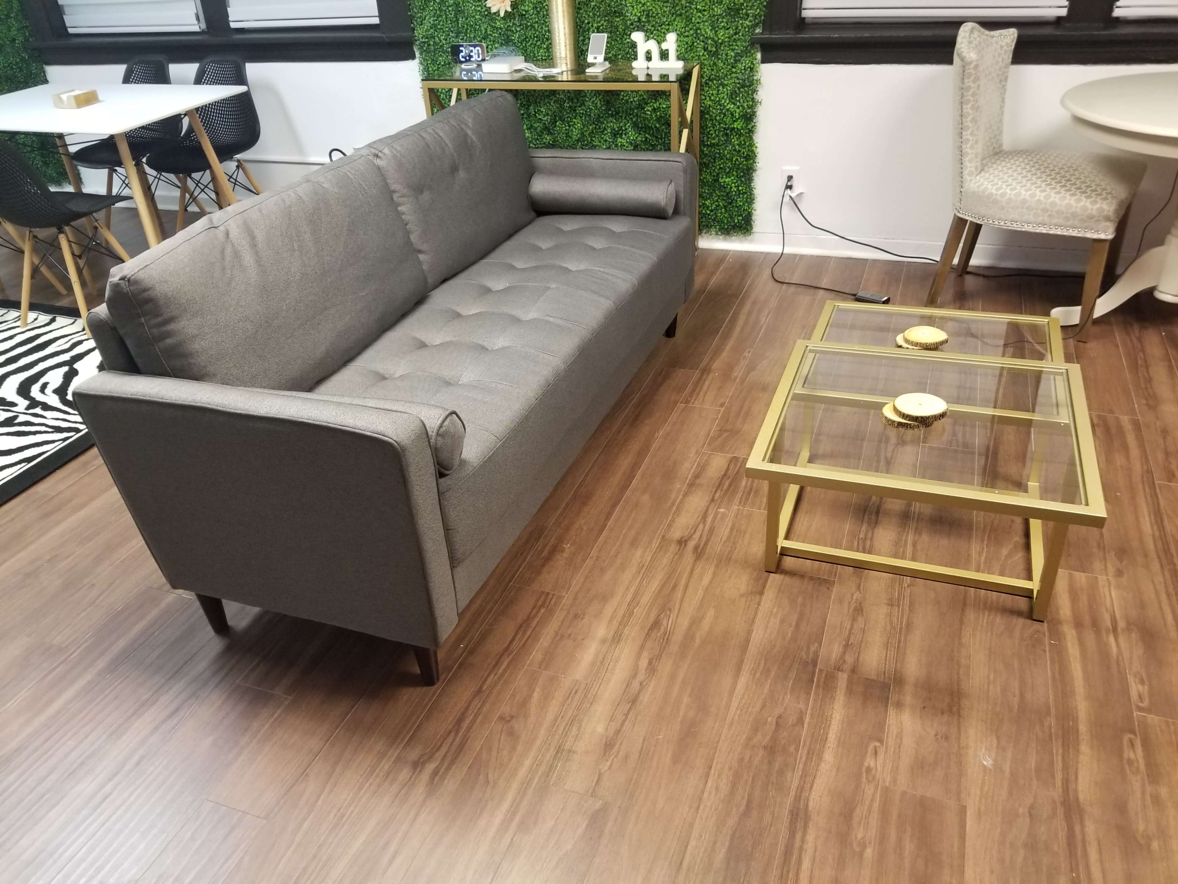 A modern gray sofa positioned beside a glass coffee table with a gold frame in a room featuring wooden flooring and green wall decor.