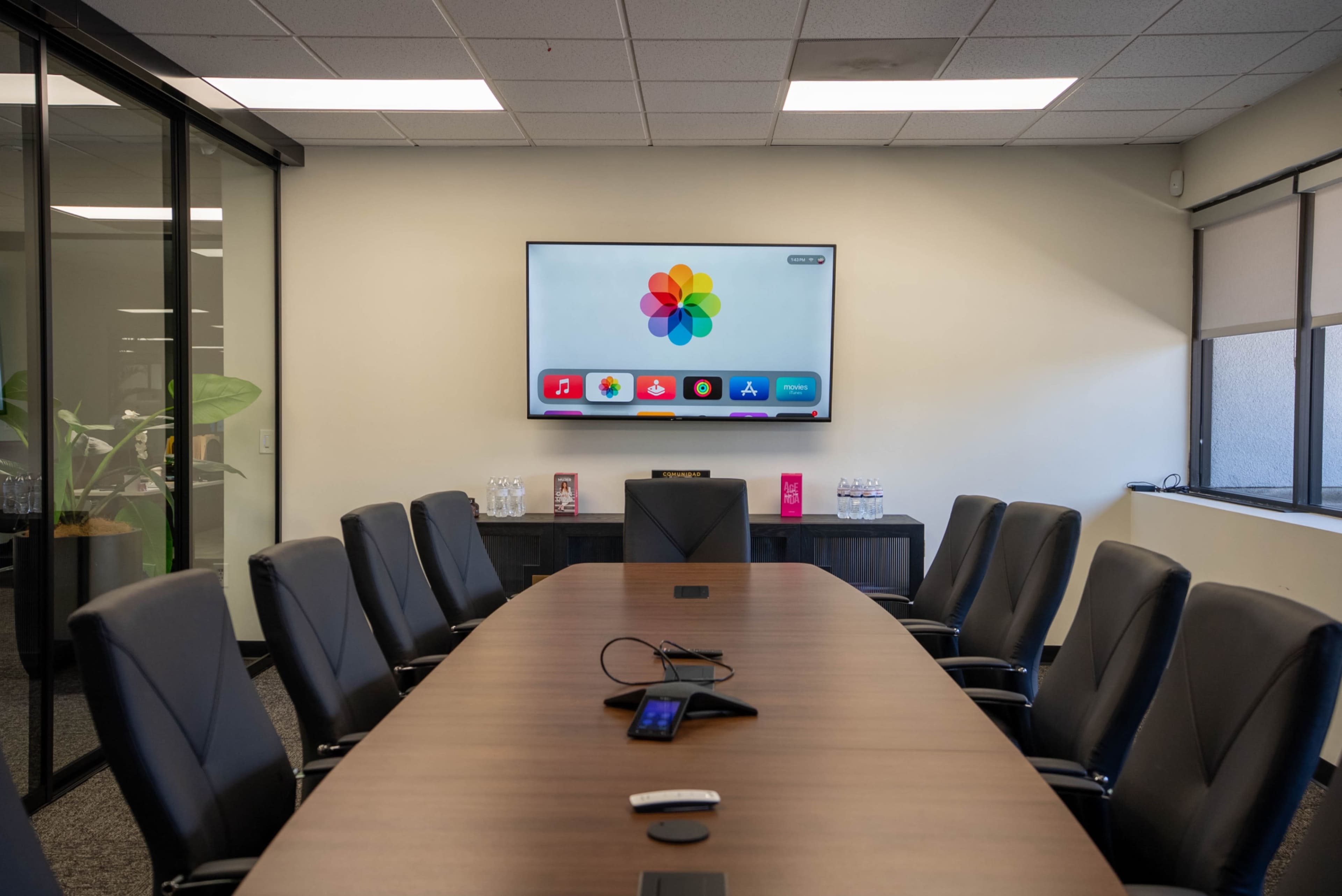 A modern conference room features a long wooden table surrounded by black leather chairs, with a wall-mounted screen displaying a colorful logo.