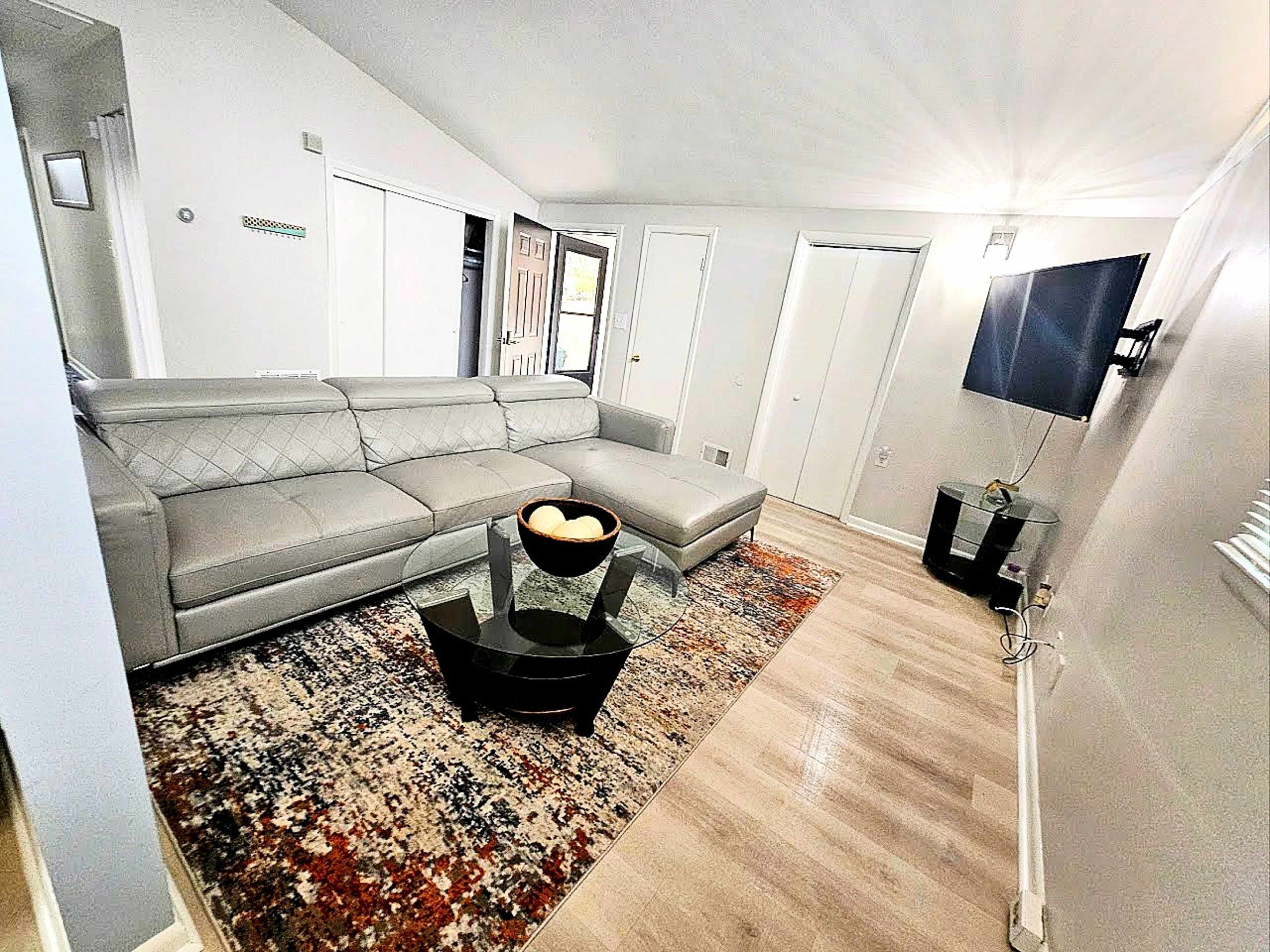 The image shows a modern living room with a gray sectional sofa, a glass coffee table on a patterned rug, a mounted TV, and light wooden flooring.