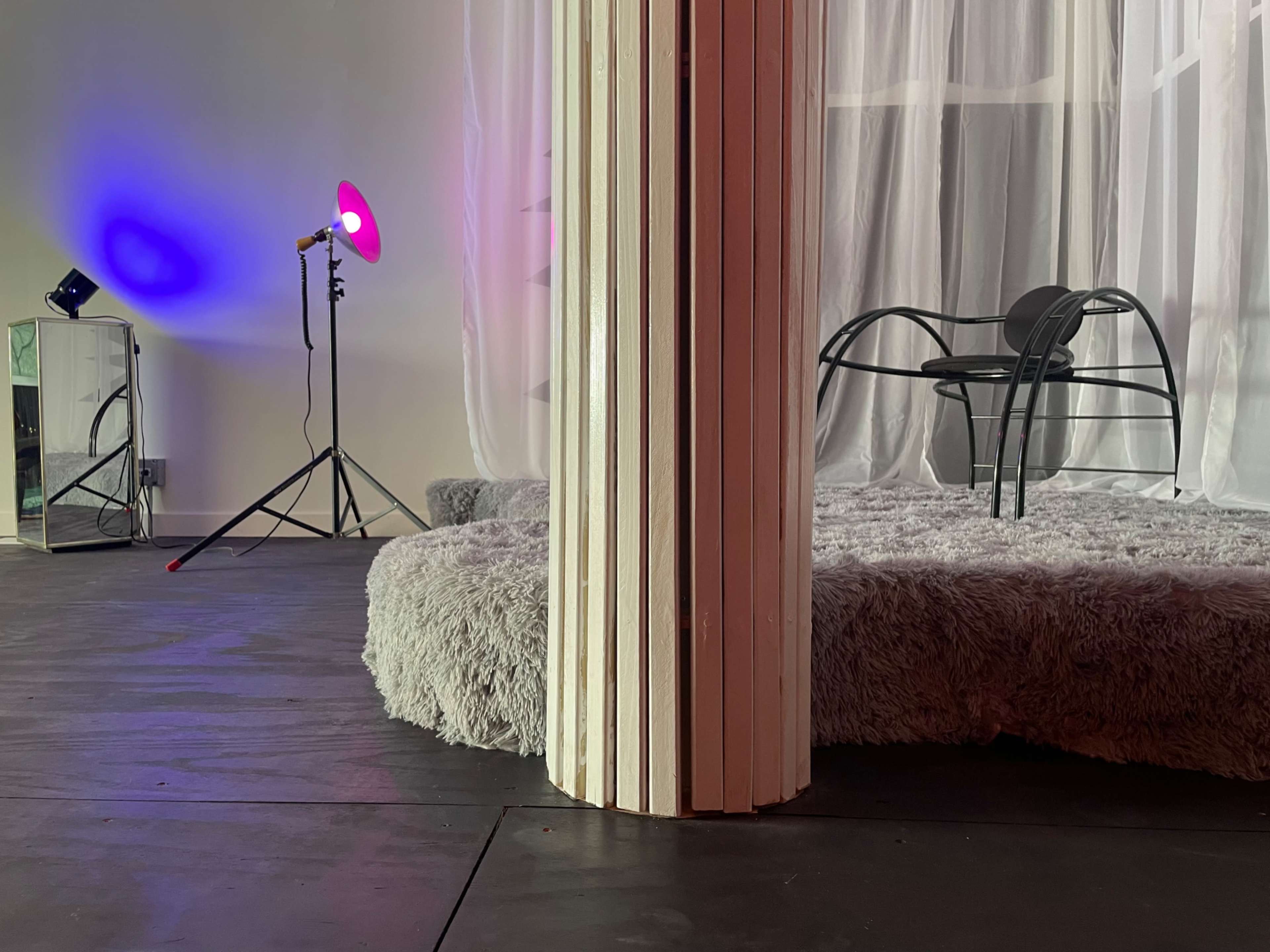 A dimly lit room with a circular, soft gray platform, a black chair, a tall standing lamp with a pink light, and a mirror reflecting the scene.