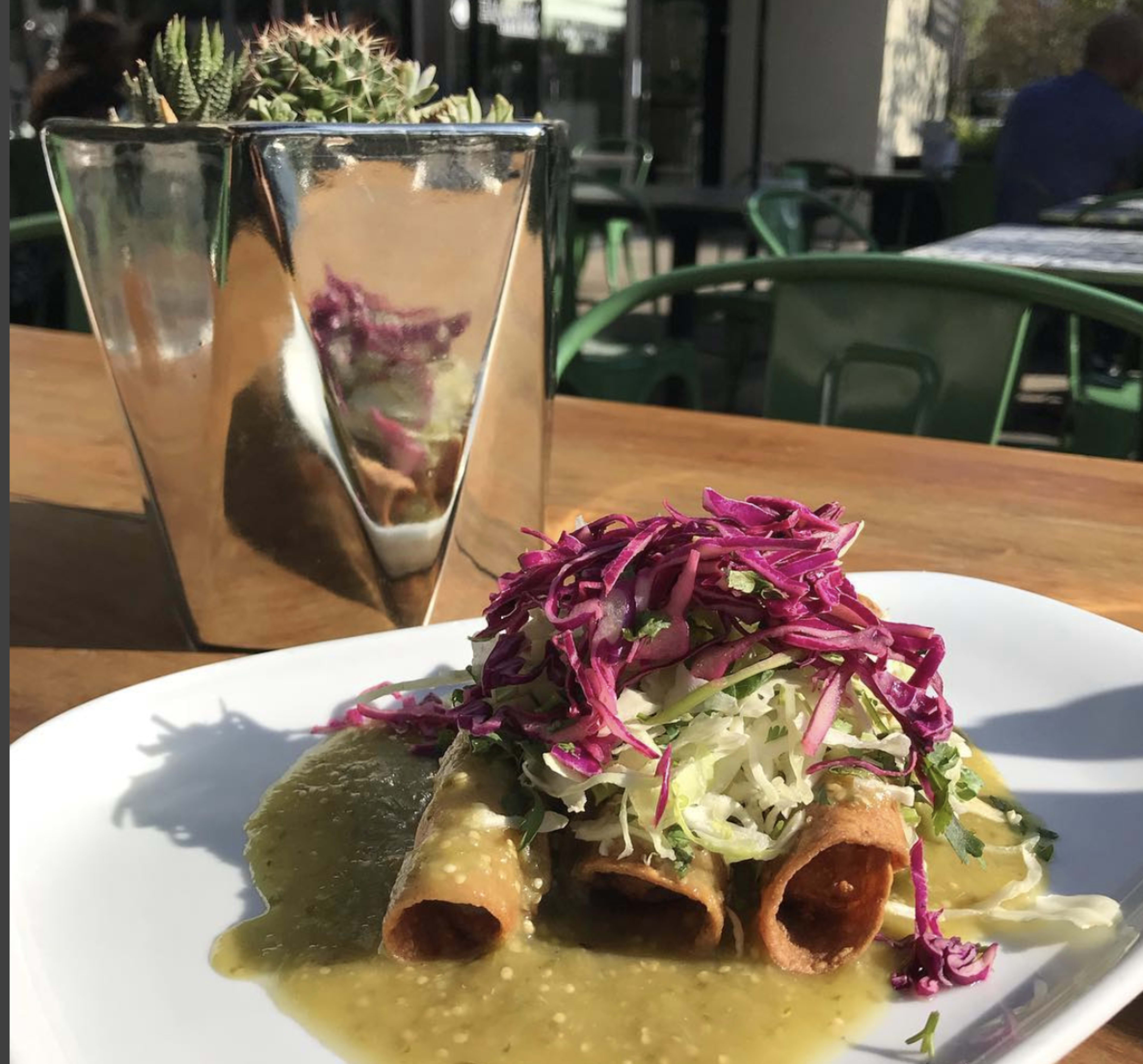 A plate of three rolled tacos topped with green sauce, shredded cabbage, and purple slaw sits on a wooden table next to a reflective metal planter with a succulent.
