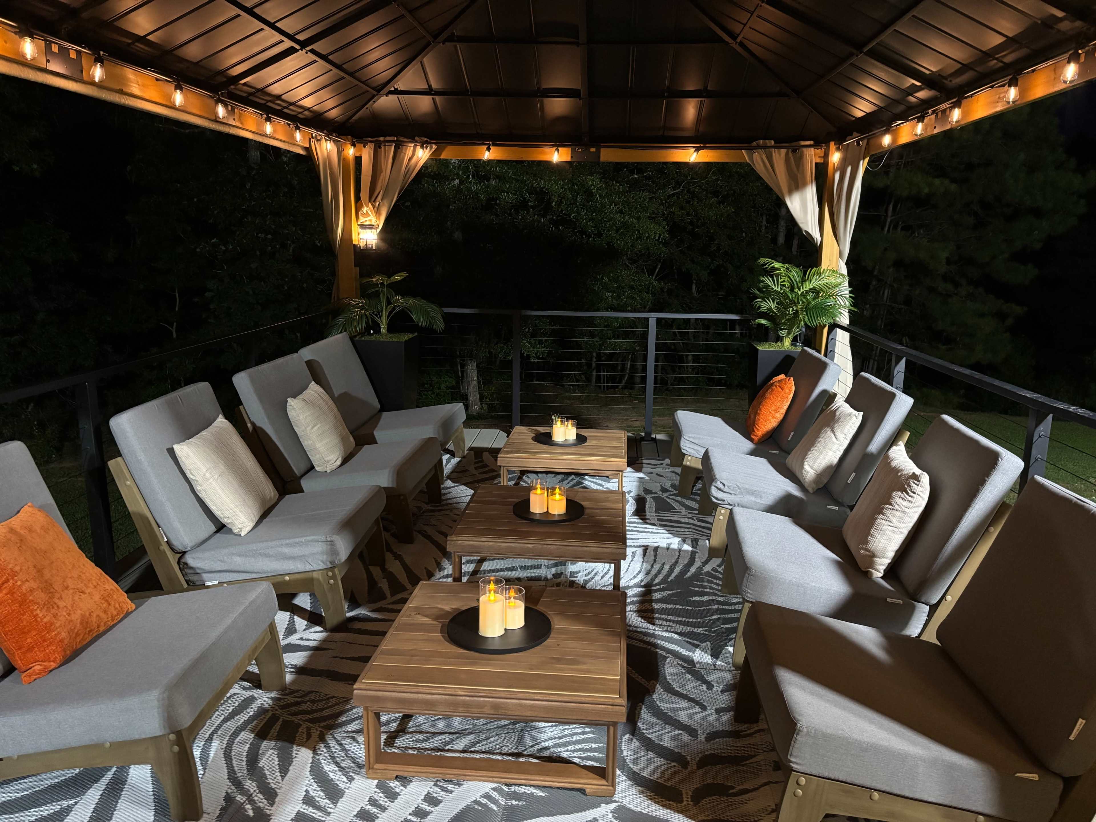 A covered outdoor seating area features several gray lounge chairs and wooden tables, illuminated by soft string lights.