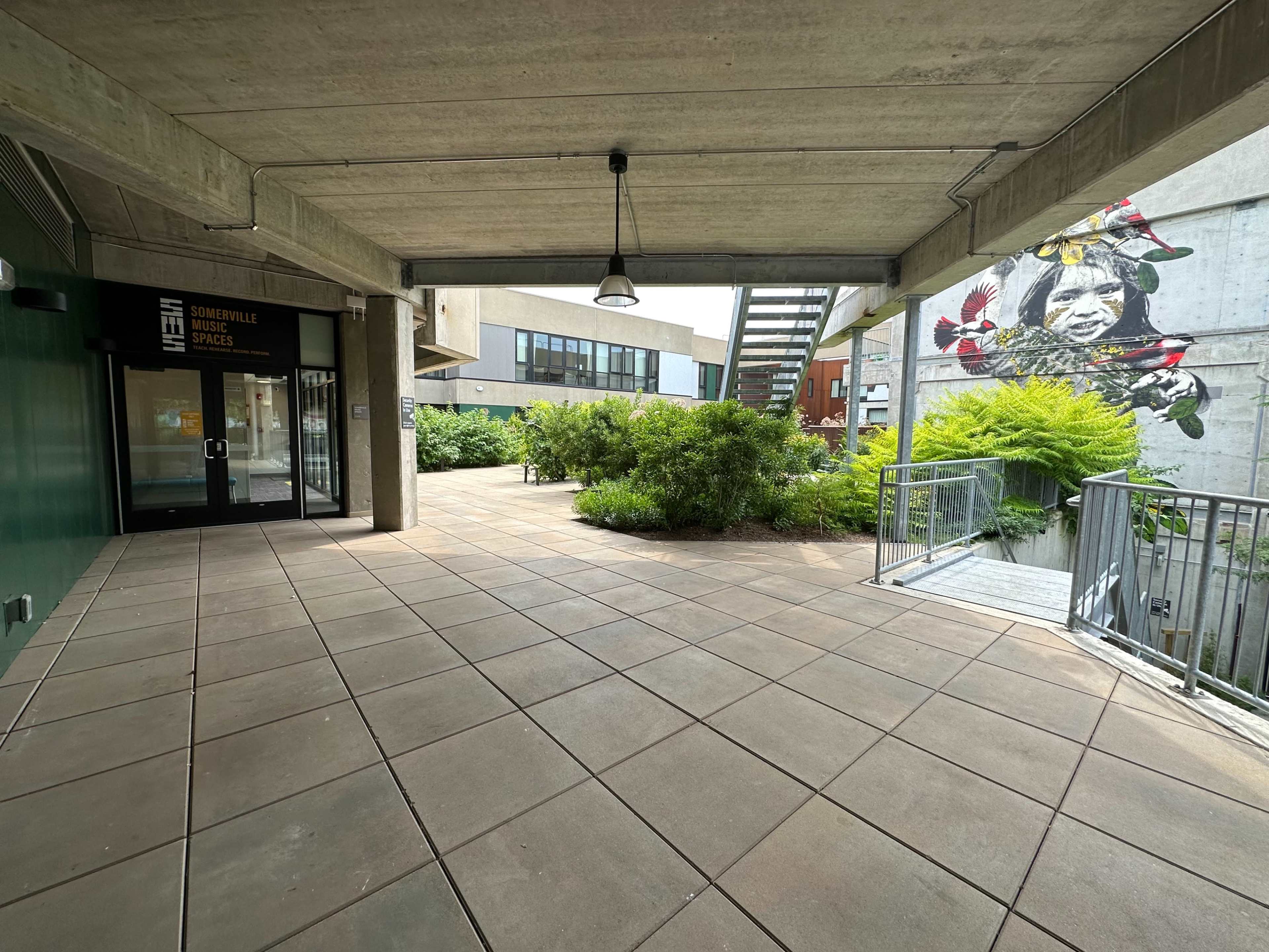 A walkway in an urban setting with concrete walls, greenery, and a mural on the right.