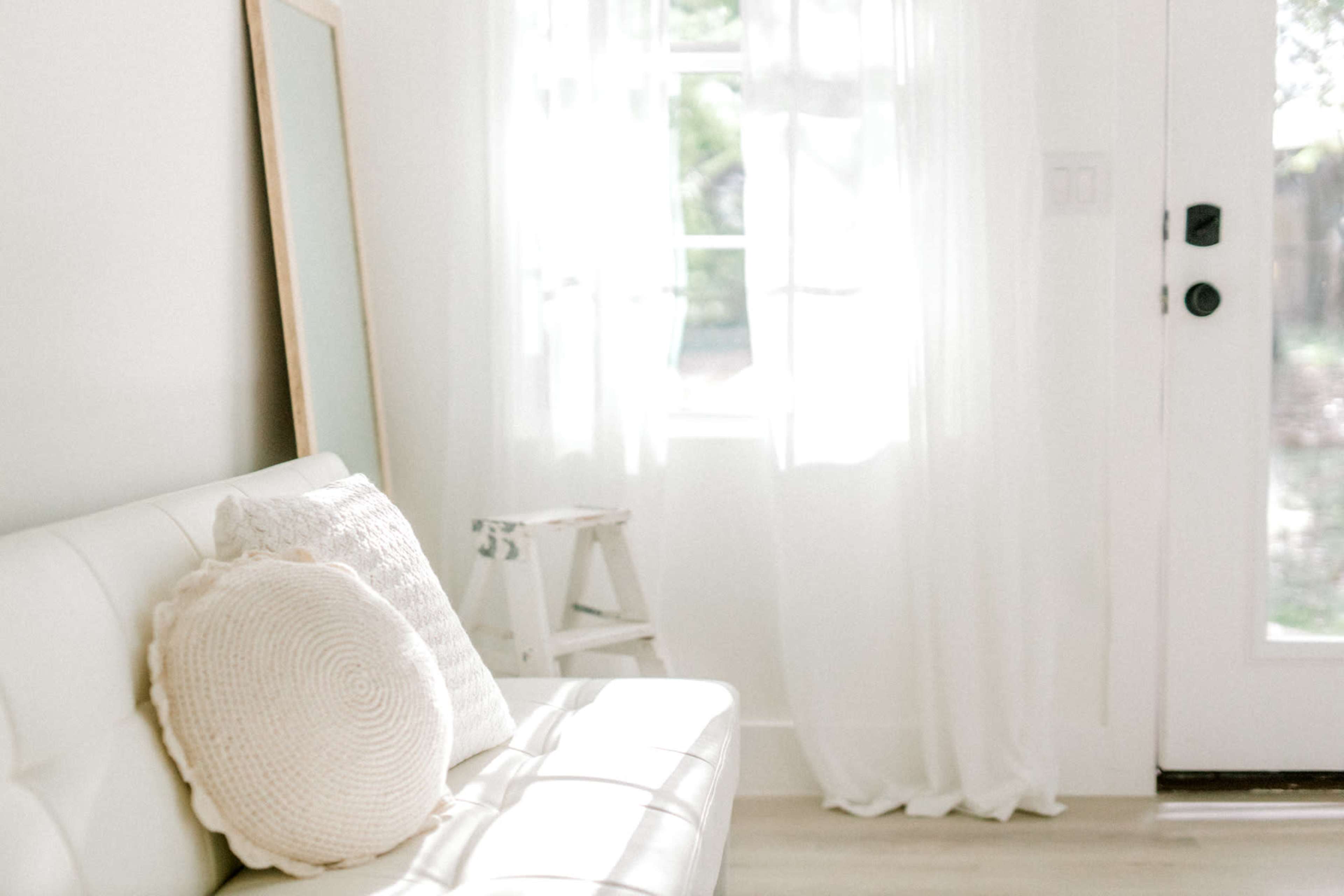 The image shows a light-colored living space featuring a white sofa with a decorative pillow, a large mirror, sheer curtains, and a door leading outside.
