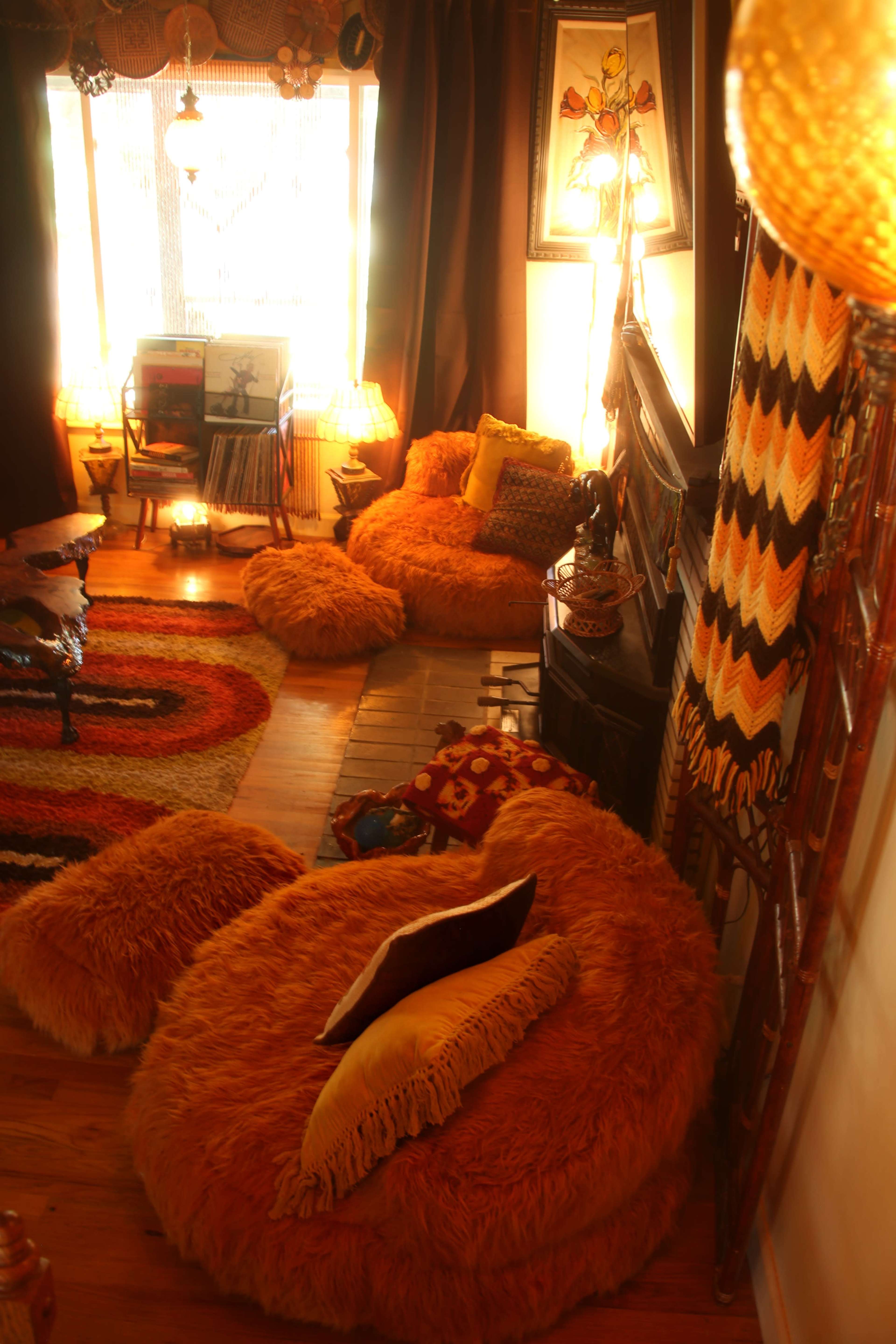 The image depicts a cozy, vintage-inspired living room with large orange bean bag chairs, colorful rugs, and warm lighting.