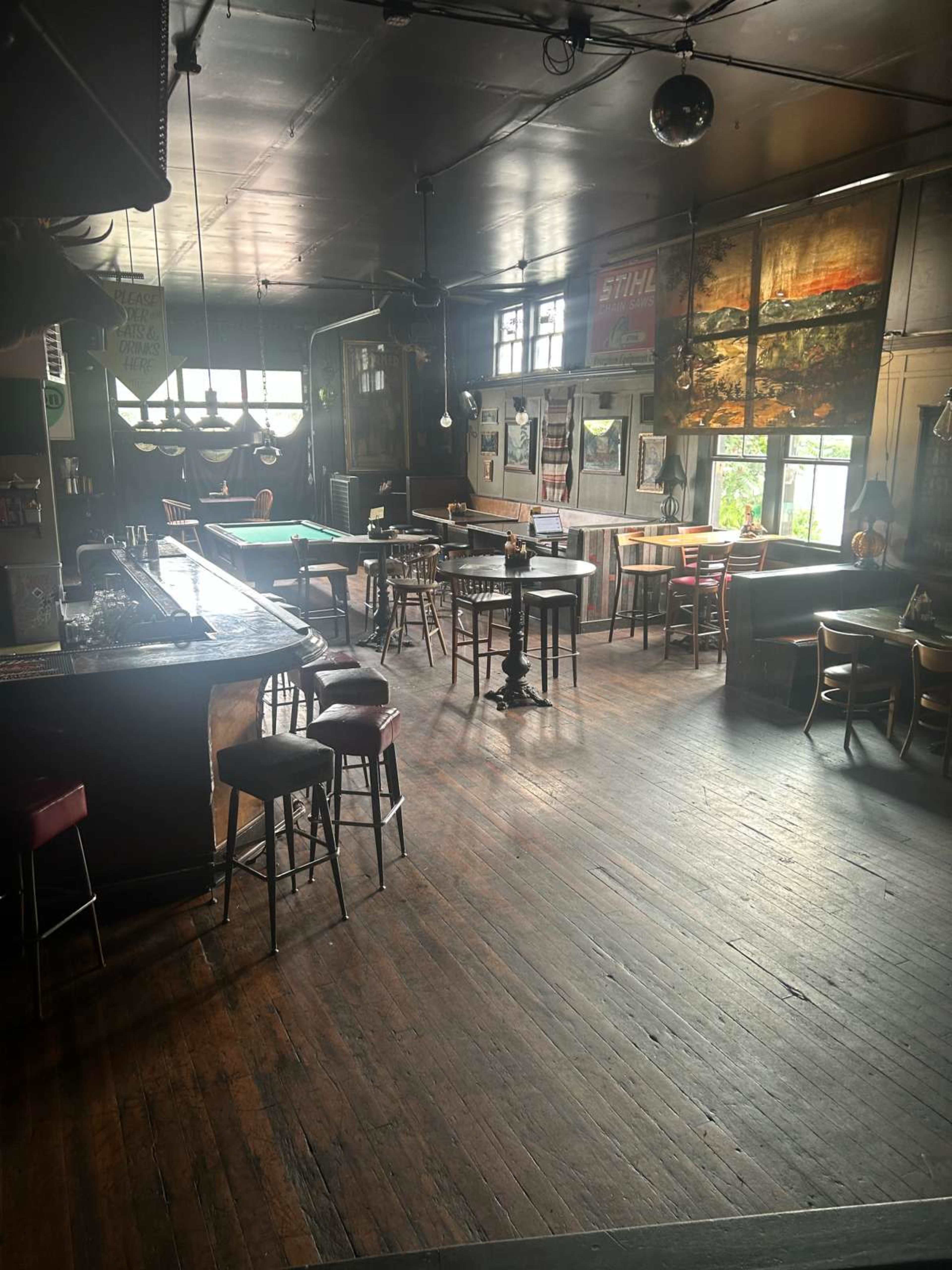 The image shows a spacious bar area with wooden floors, various tables and chairs, a pool table, and large windows letting in natural light.