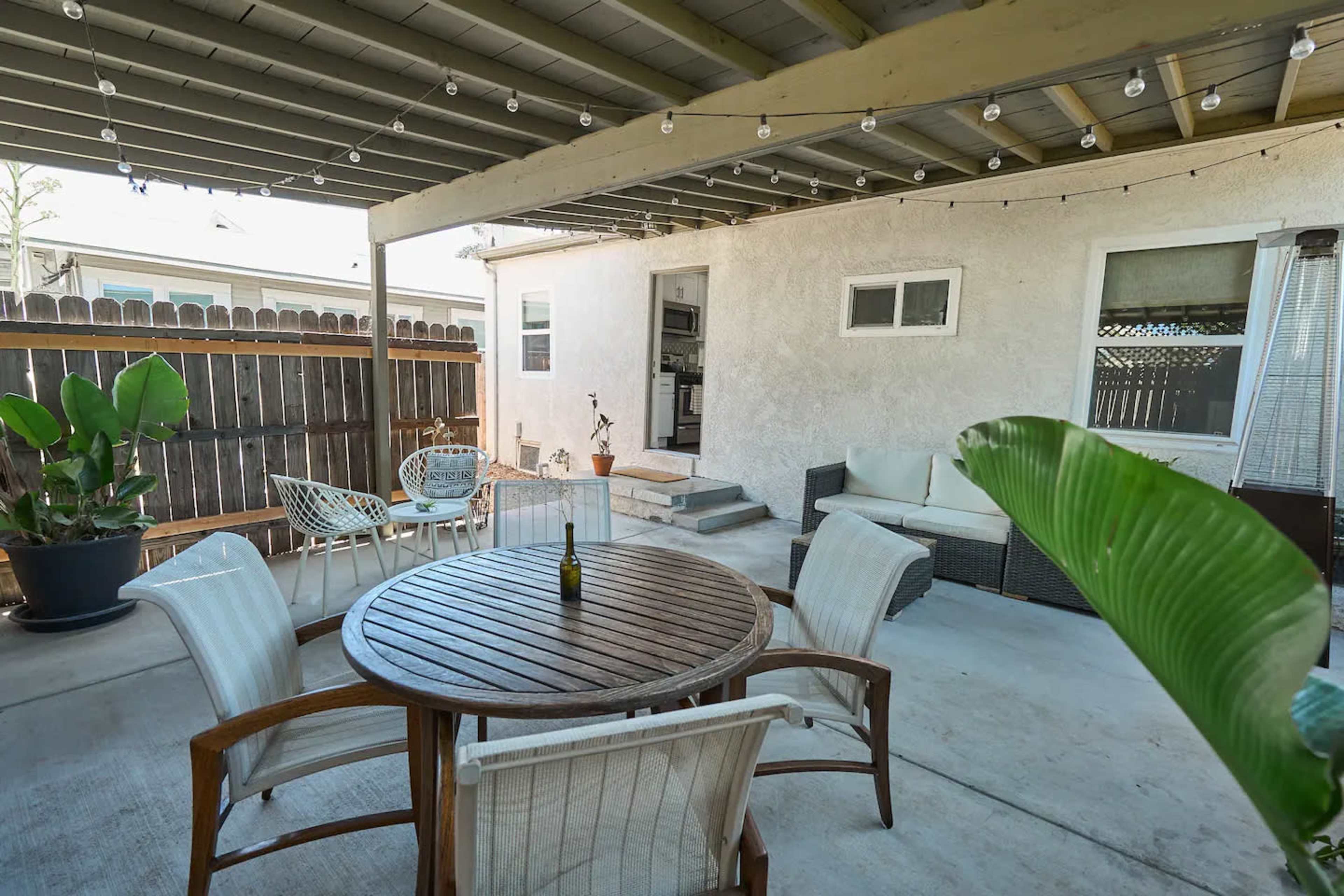 The image shows a covered outdoor patio with a round wooden table surrounded by chairs, a small potted plant, and a sofa area against a wall.