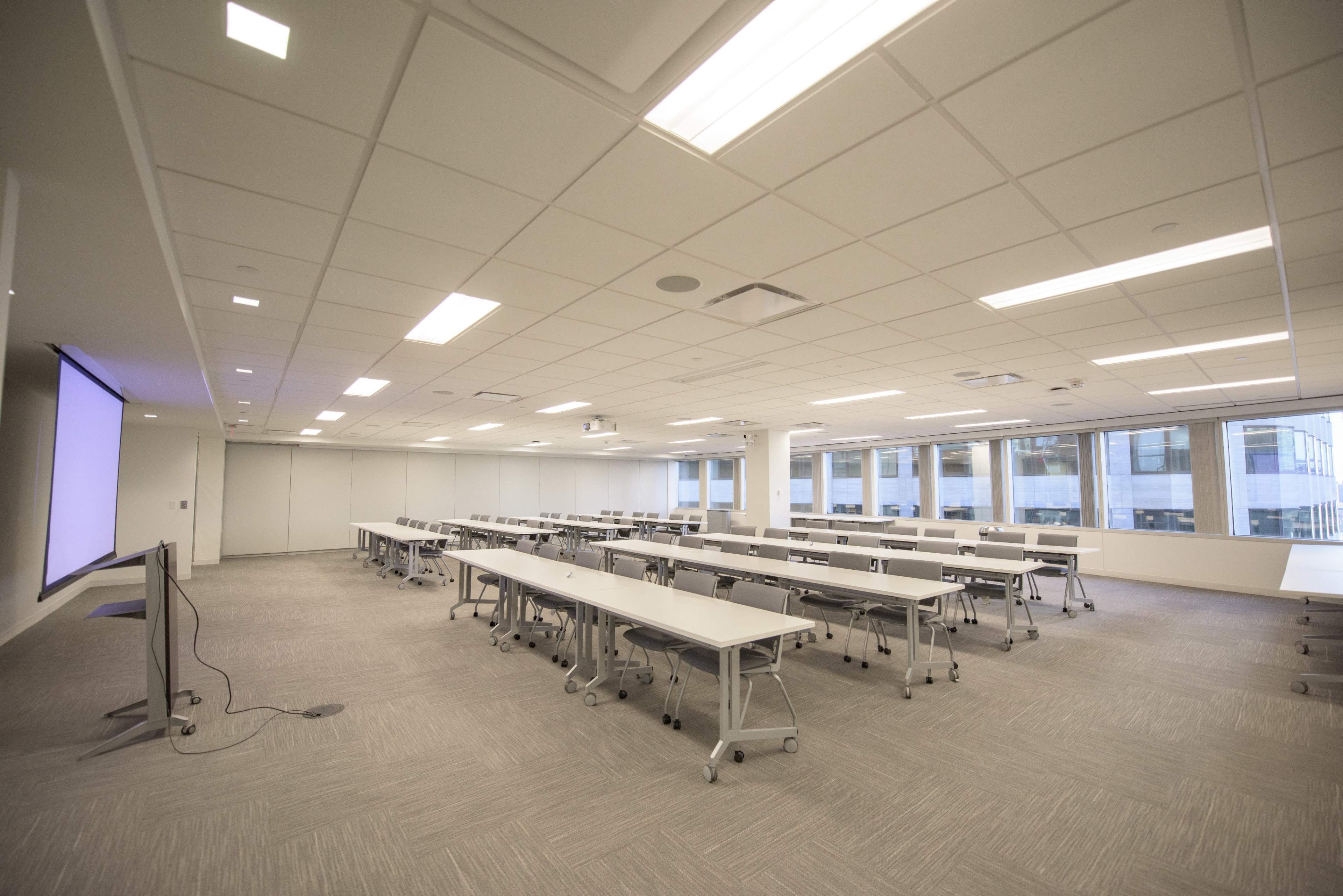 A bright, spacious conference room features multiple rows of tables and chairs, with large windows allowing natural light to enter.
