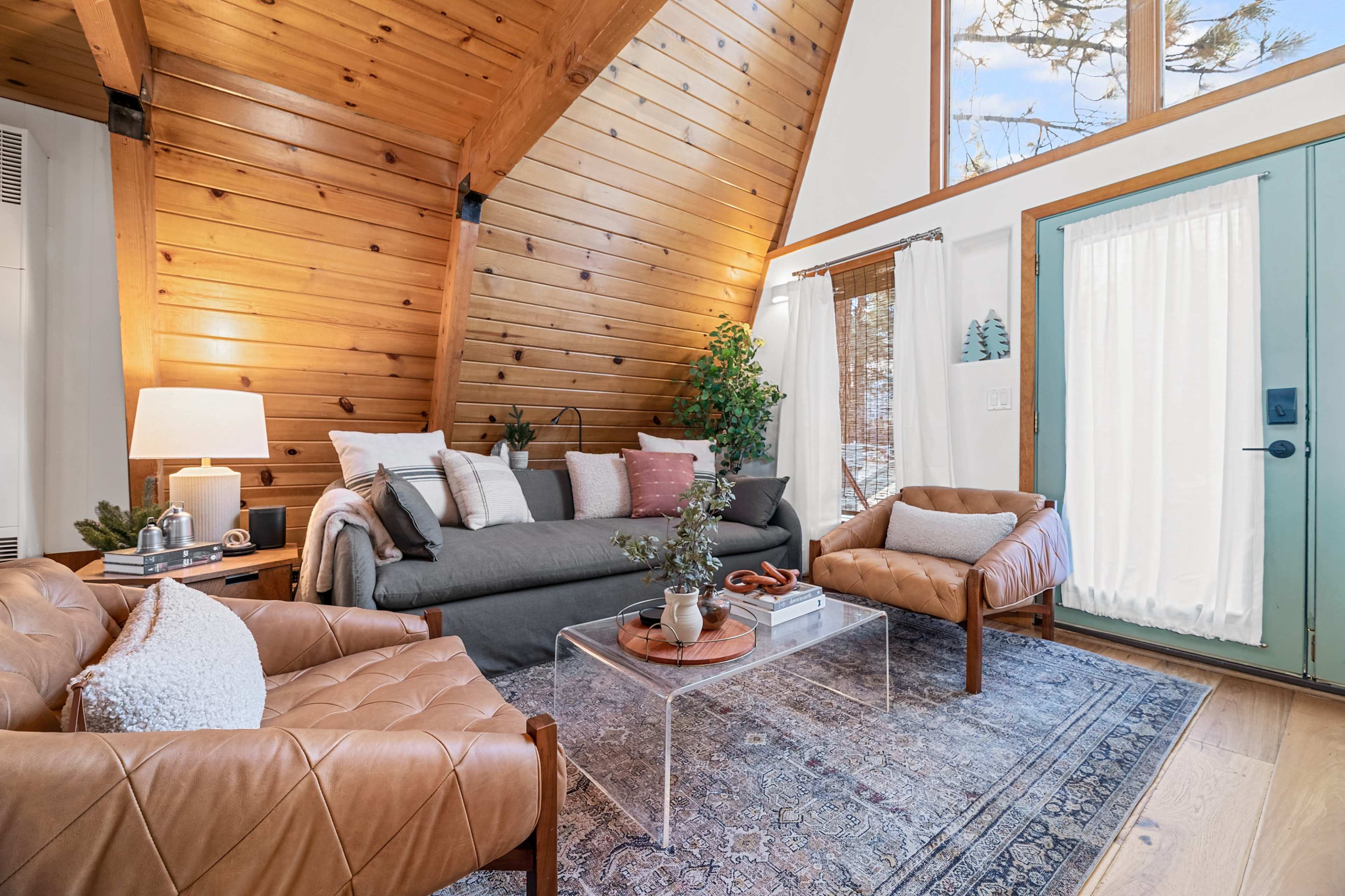 A cozy living room with a high wooden ceiling, a grey sofa, leather chairs, and a glass coffee table on a patterned rug, complemented by a door that opens to the outside.