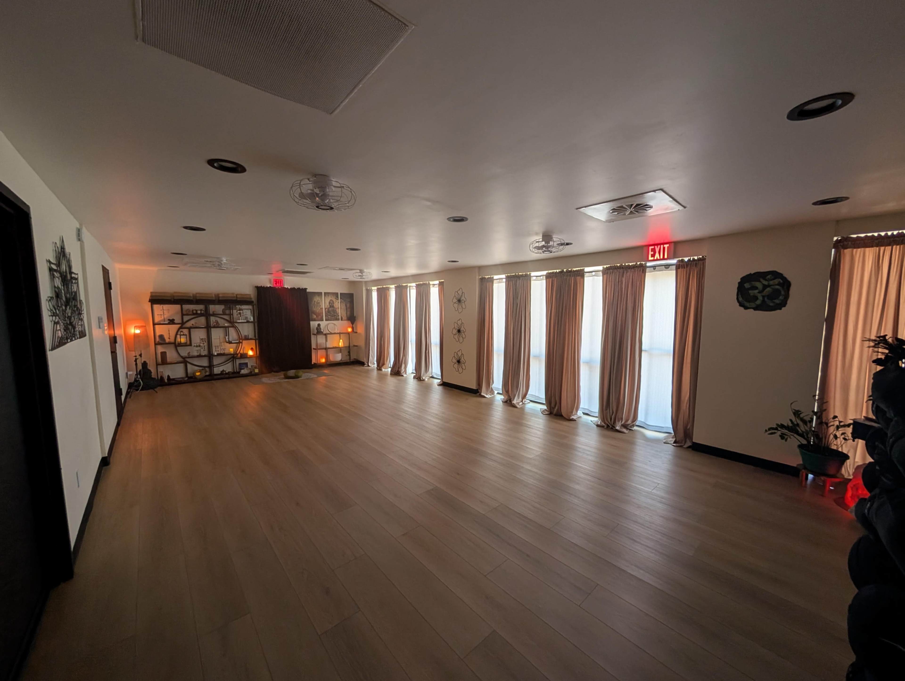 The image shows a spacious room with wooden flooring, large windows covered by light curtains, and decorative elements along the walls.
