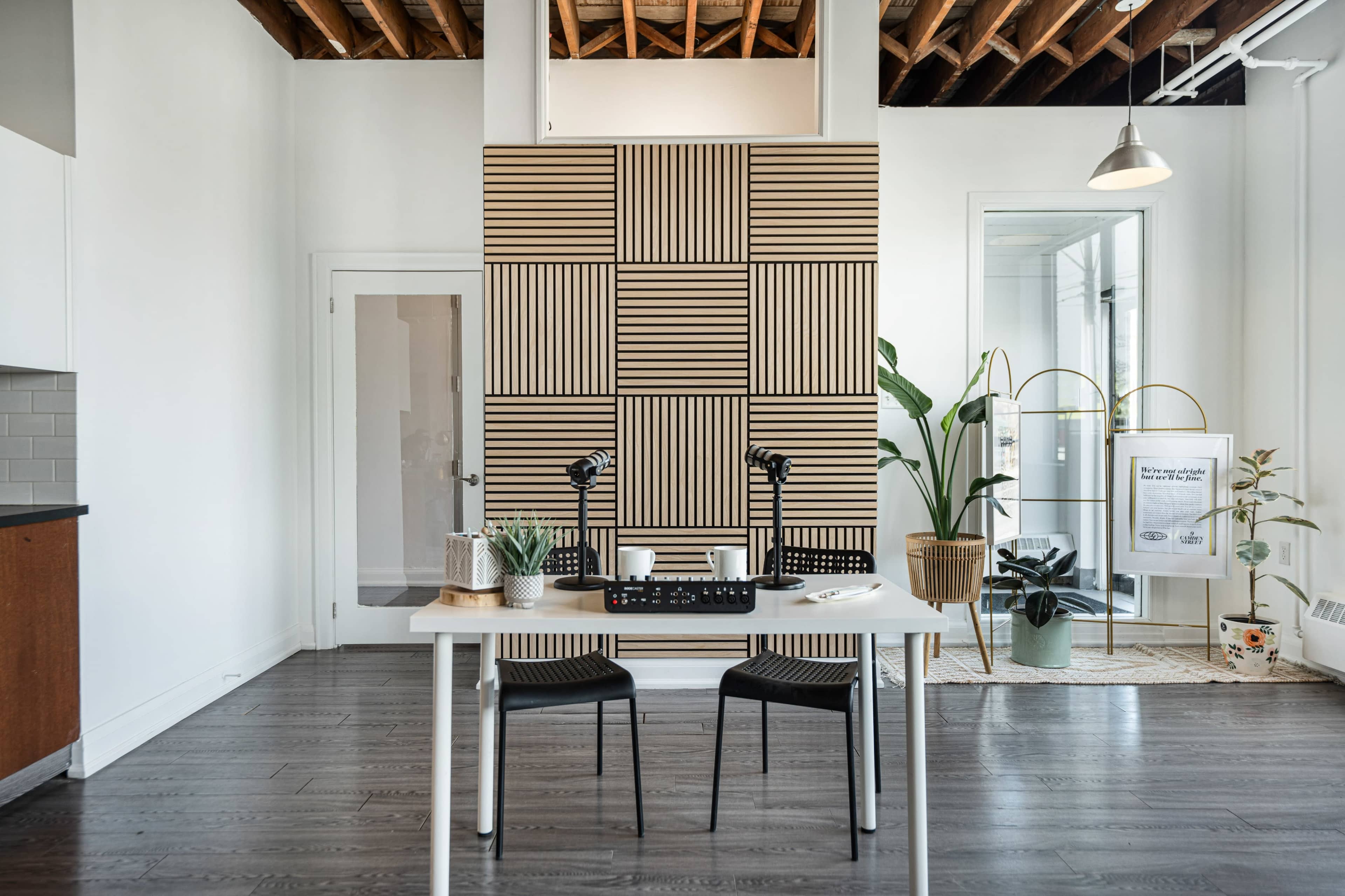 A minimalist workspace features a table with two black chairs, a geometric wall design, and indoor plants against a backdrop of large windows and wood ceiling beams.