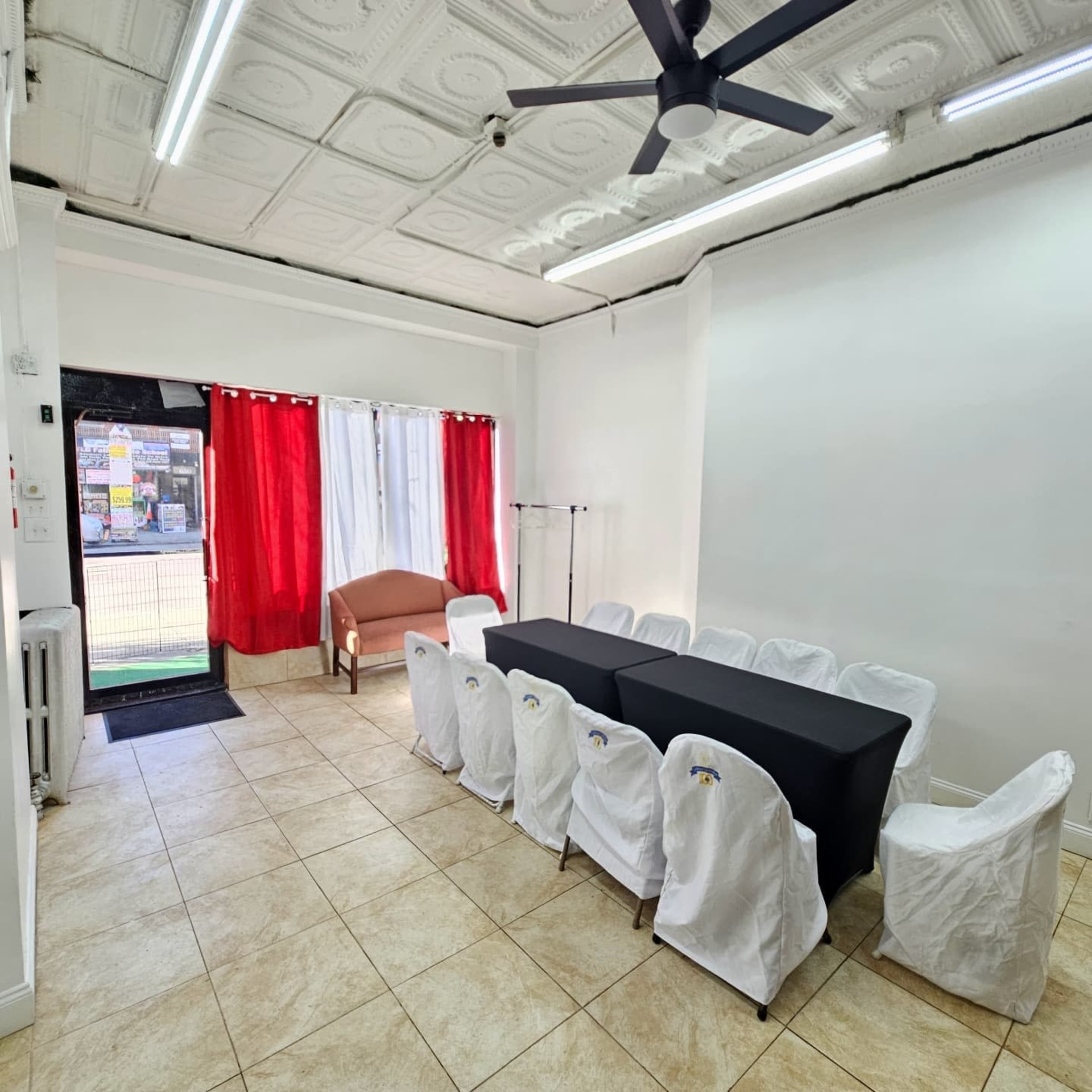 The image shows a well-lit room with a black table surrounded by white-covered chairs, red curtains at the windows, and a beige couch near the entrance.