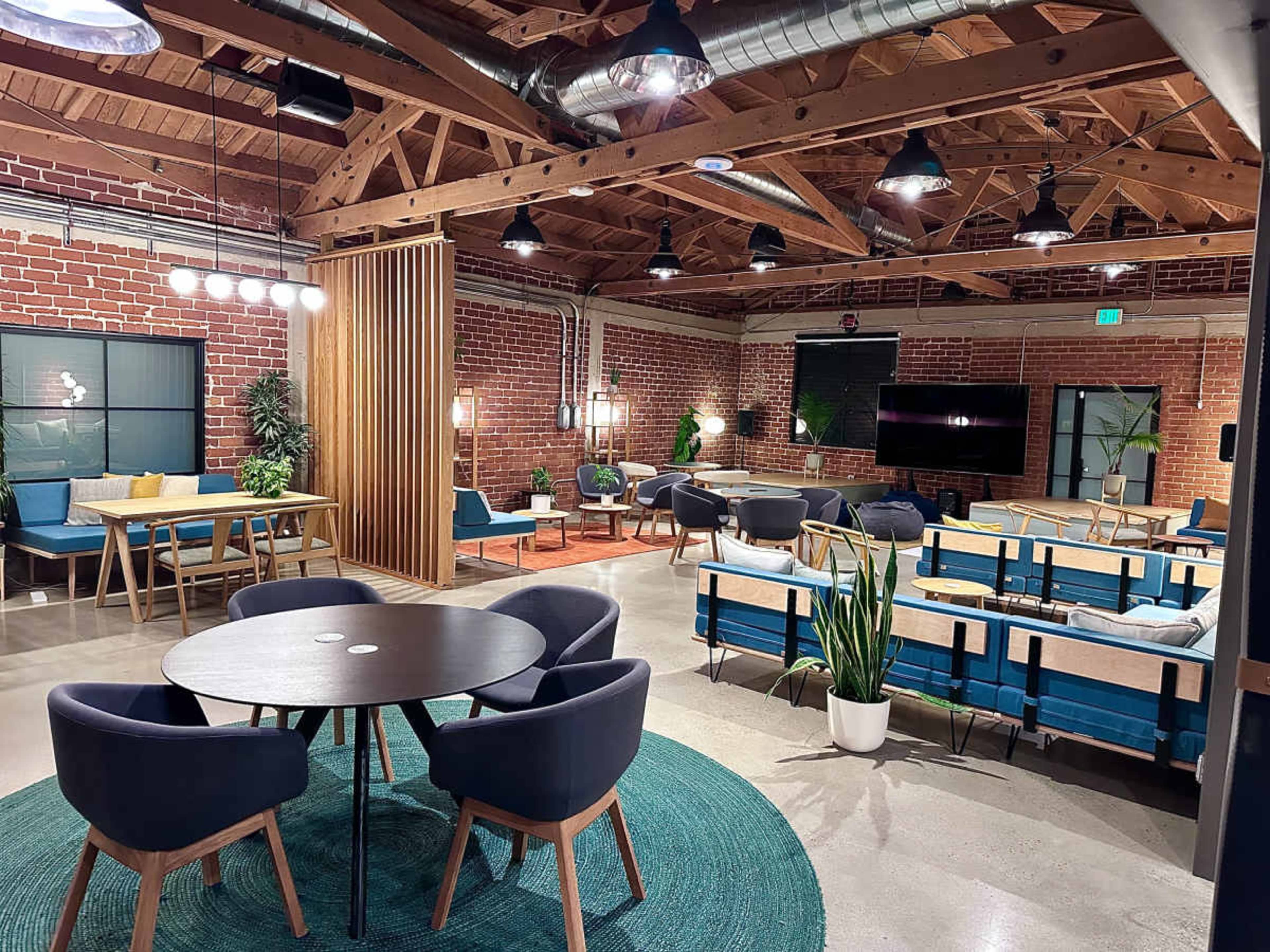 The image shows a modern, open-concept lounge area with wooden beams, brick walls, and various seating arrangements.