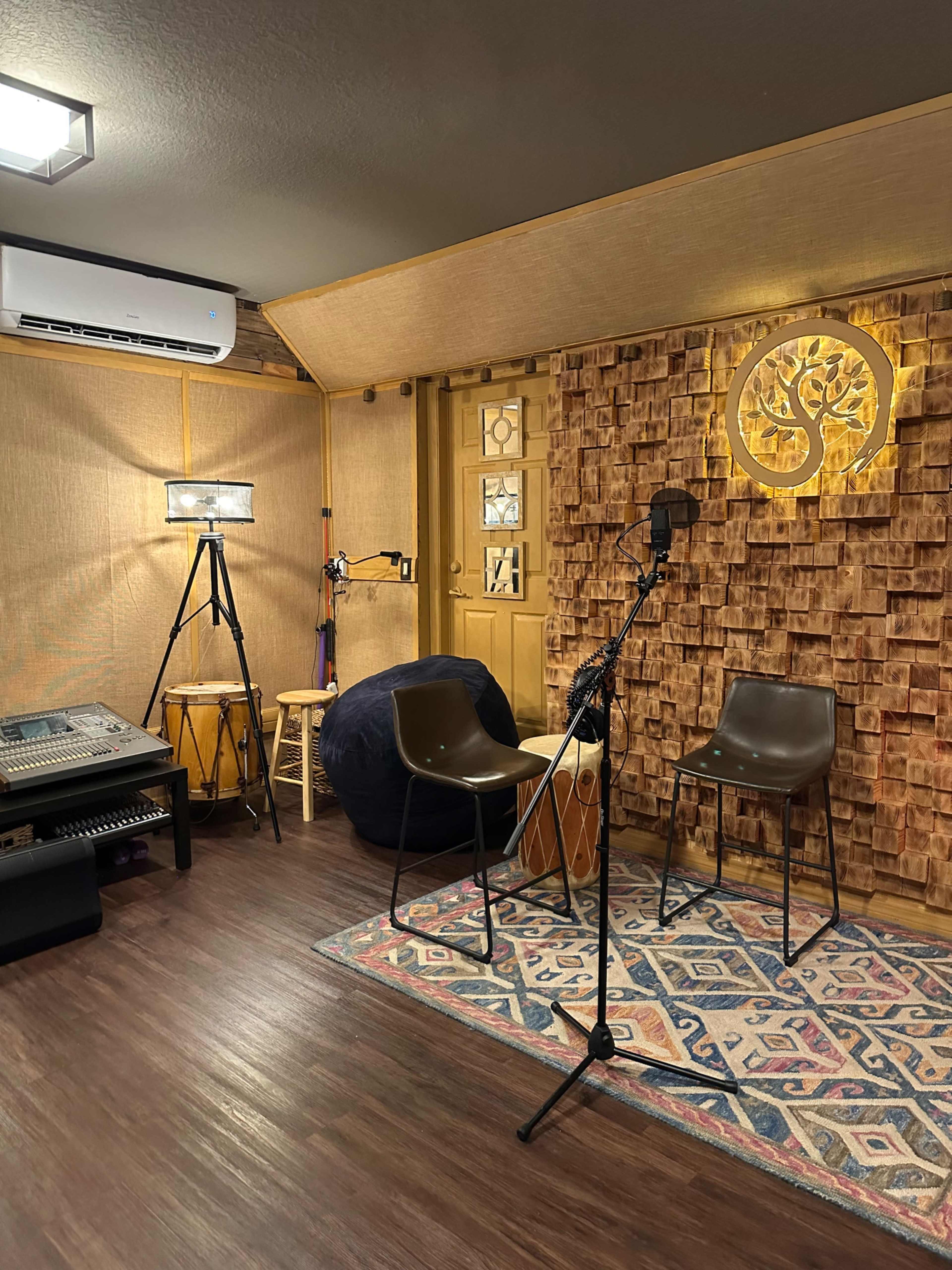 A small recording studio featuring two chairs, a microphone on a stand, a camera on a tripod, and various musical instruments against a textured wall.