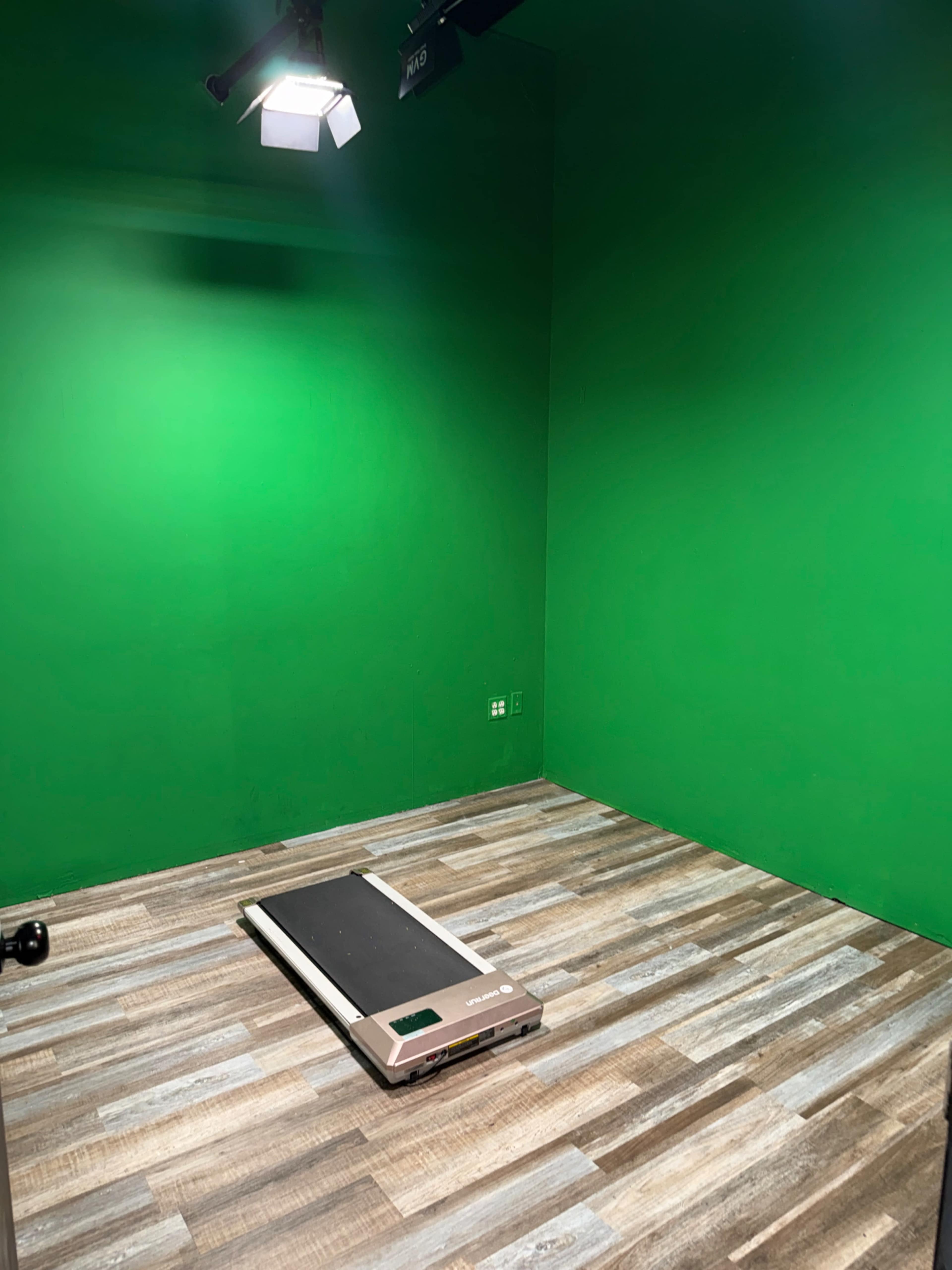 The image shows a small room with a bright green wall, wooden flooring, and a treadmill placed in the center under a light fixture.