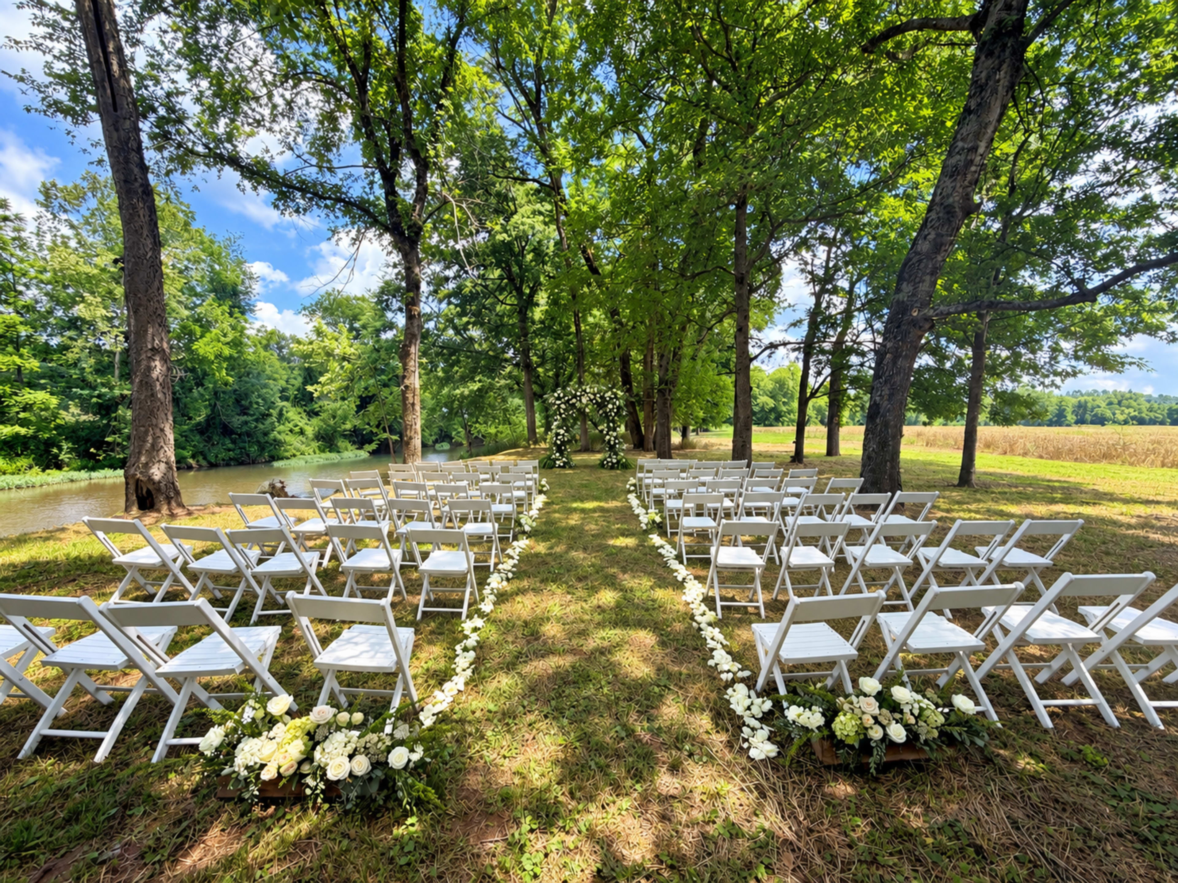 A wedding ceremony setup features rows of white chairs arranged on a grassy space under trees, with a floral arch at the end facing a serene backdrop of water and fields.