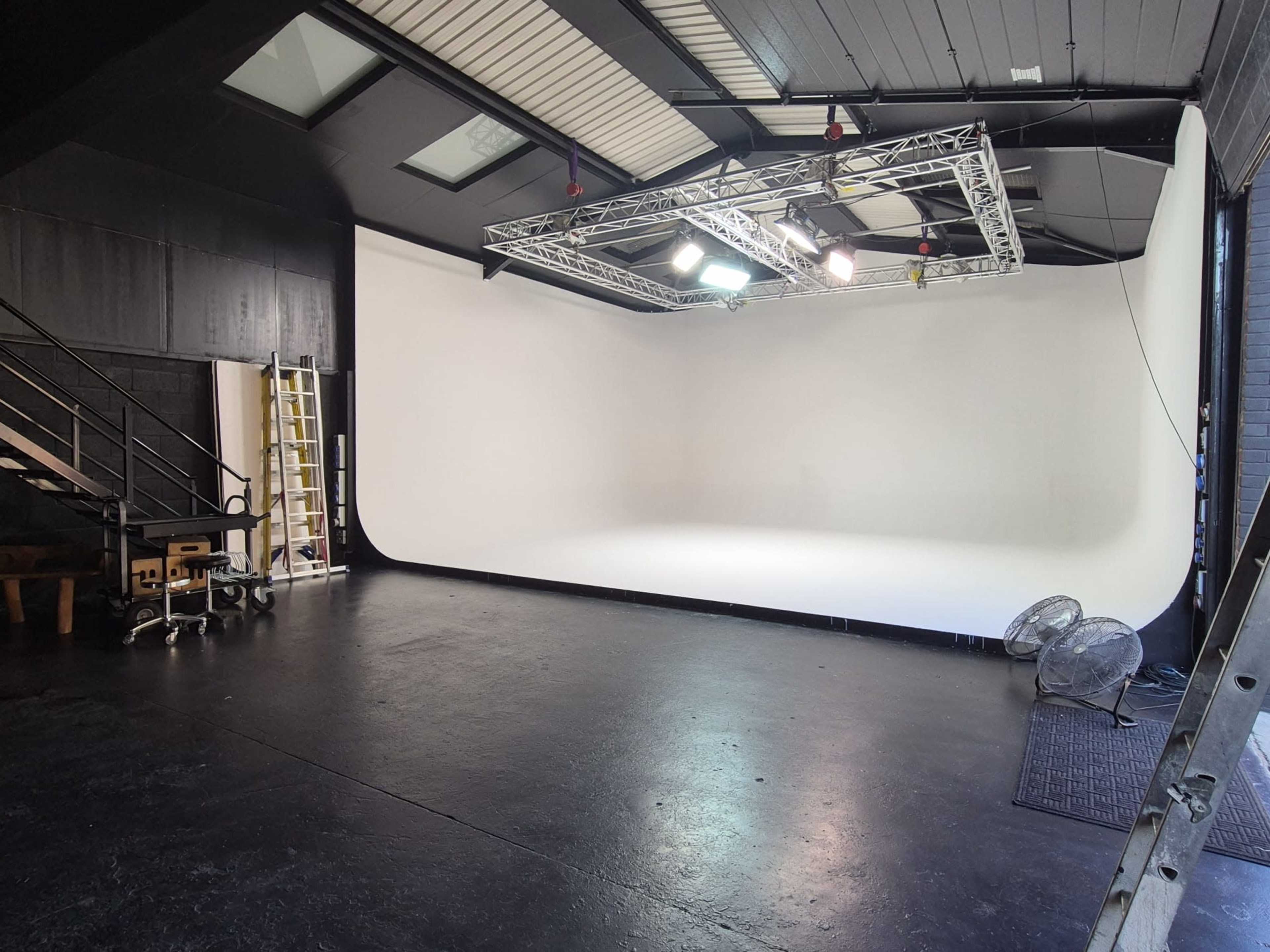 The image shows a spacious, empty studio with a seamless white backdrop, overhead lighting rigs, and a staircase in the background.