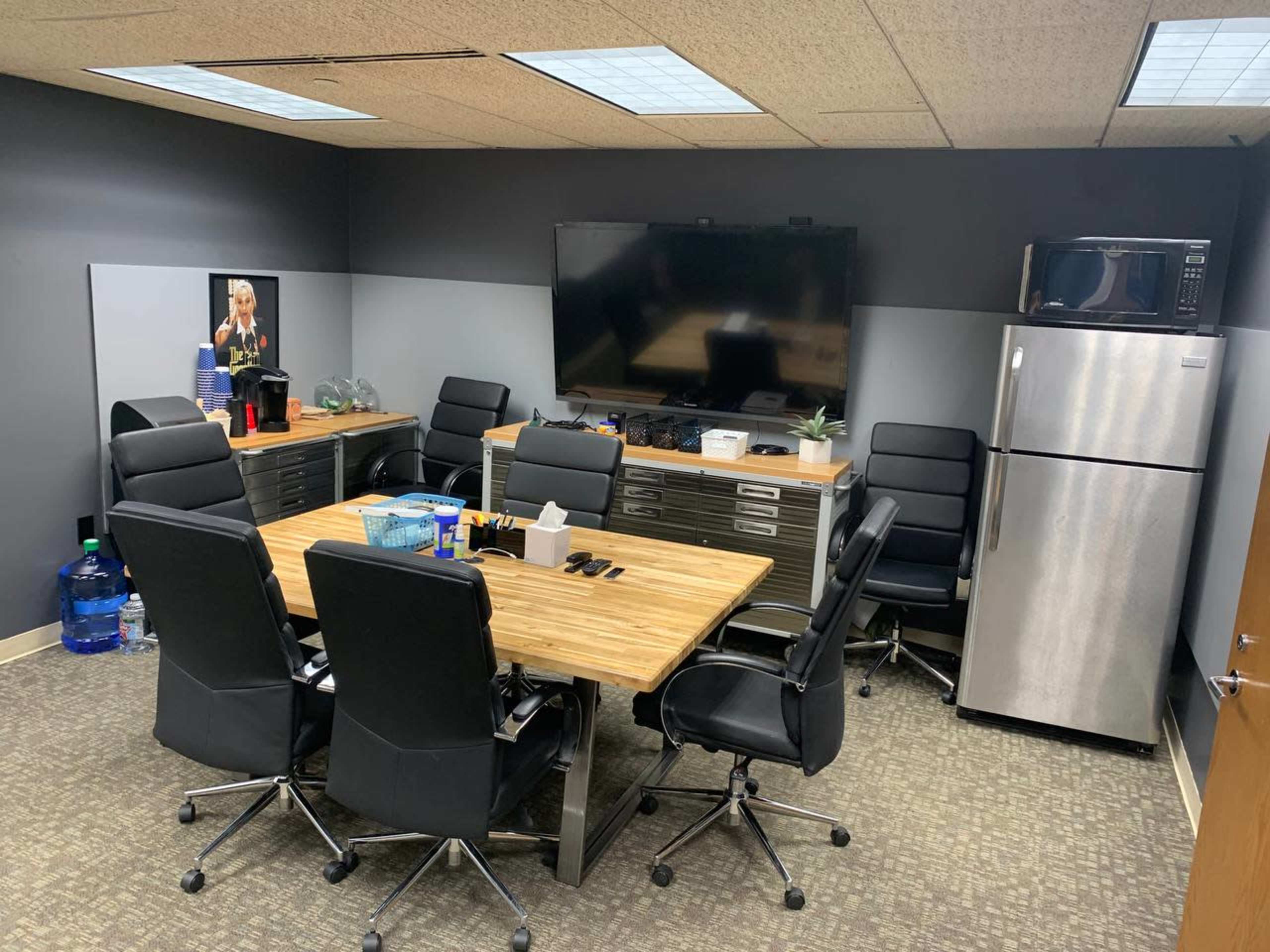 A conference room features a large wooden table surrounded by black chairs, a television on the wall, and a kitchen area with a microwave and a refrigerator.