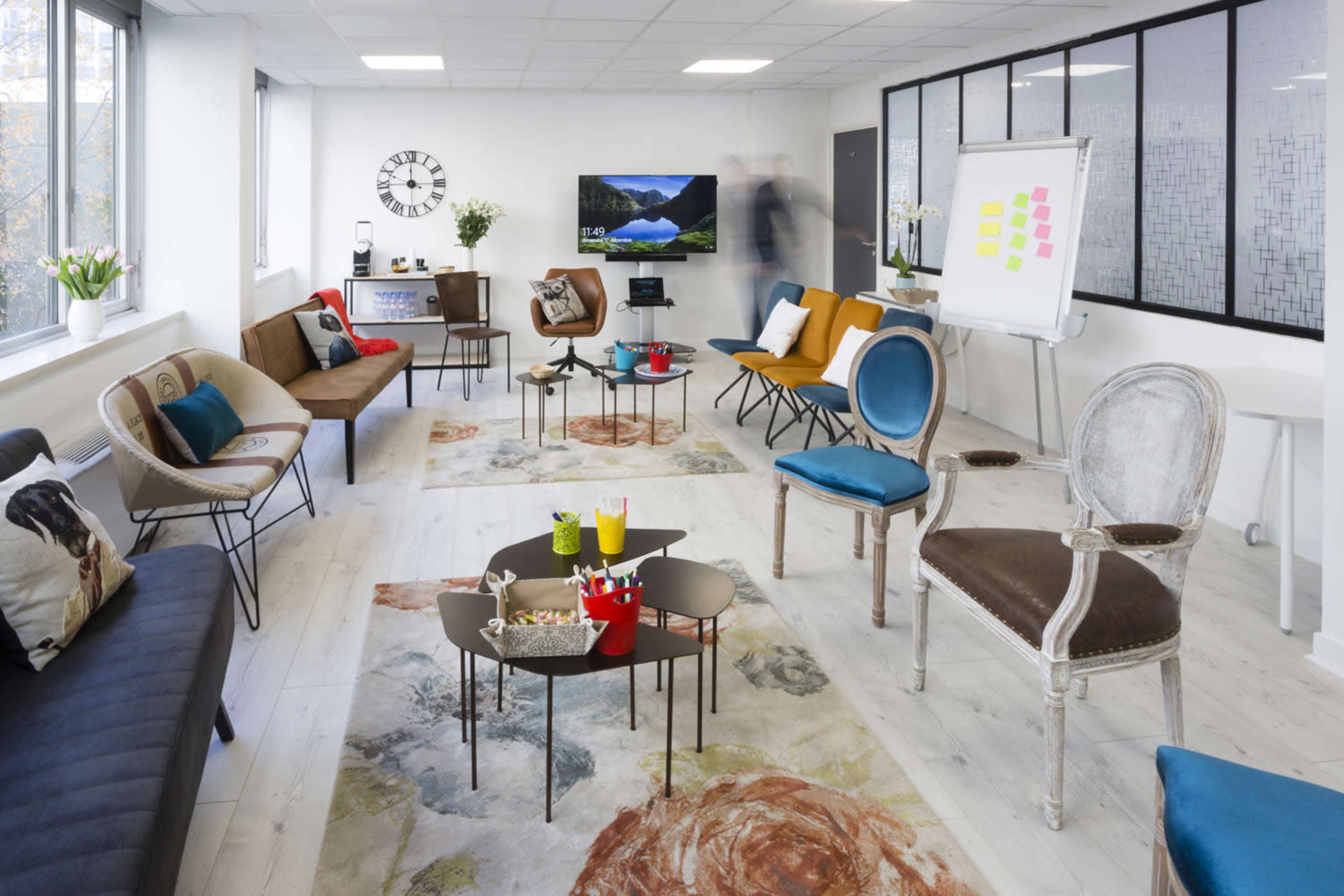 A modern, spacious meeting room filled with various seating options and tables, featuring large windows and a presentation area with a whiteboard.