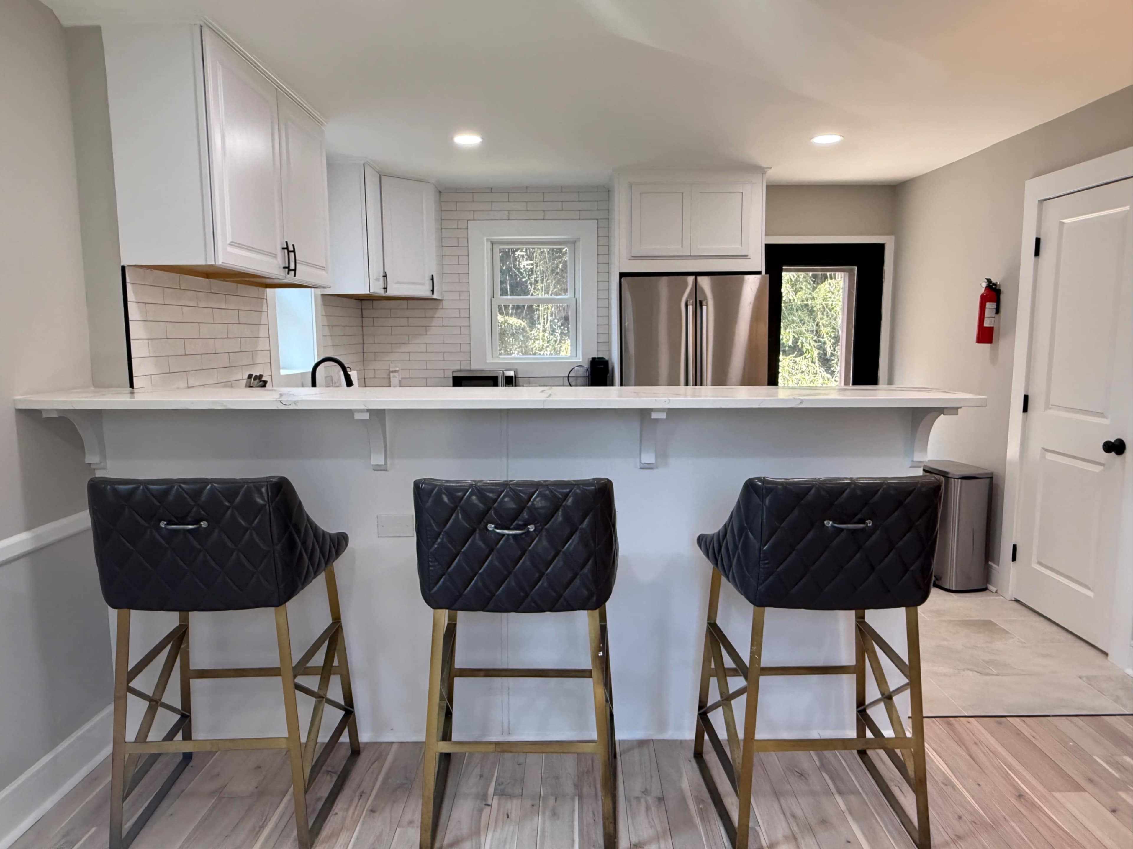 The image shows a modern kitchen with a white cabinetry, a bar area featuring three black upholstered stools, and a stainless steel refrigerator in the background.