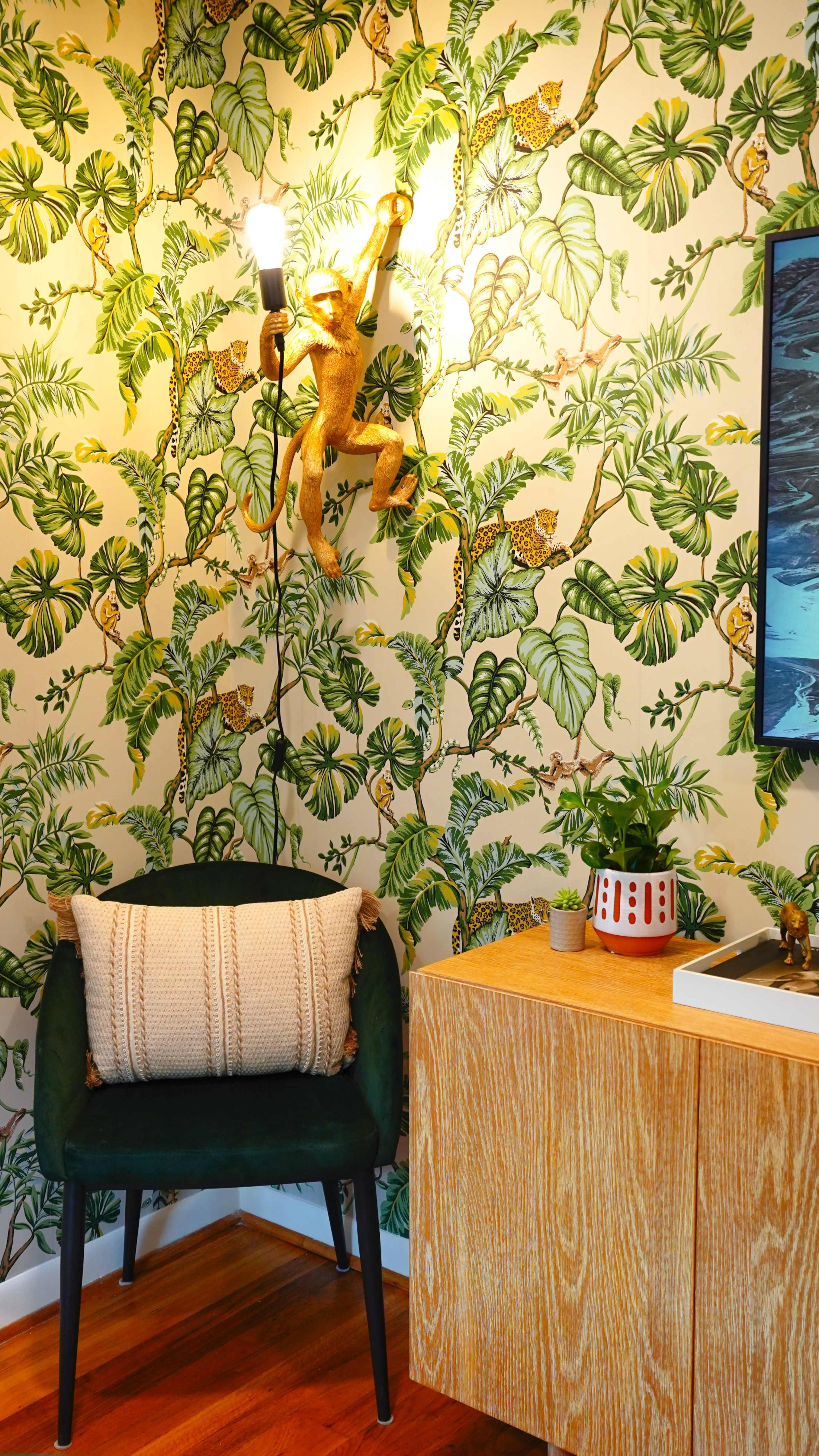 The image features a corner of a room with a green upholstered chair, a wooden side table, and wallpaper adorned with leafy patterns and animal motifs.