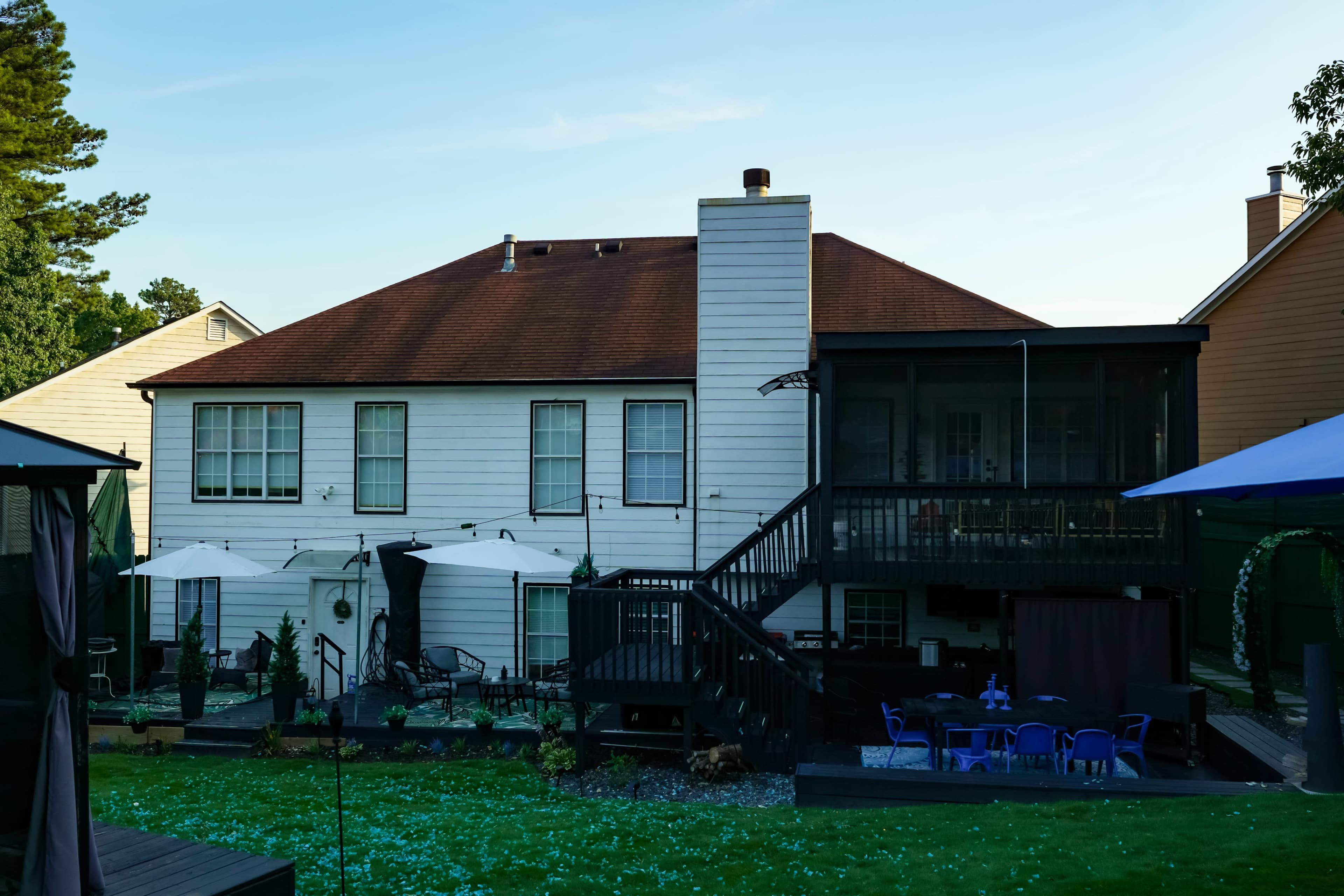 The image shows the back of a two-story house with a spacious deck, outdoor furniture, and a landscaped yard.
