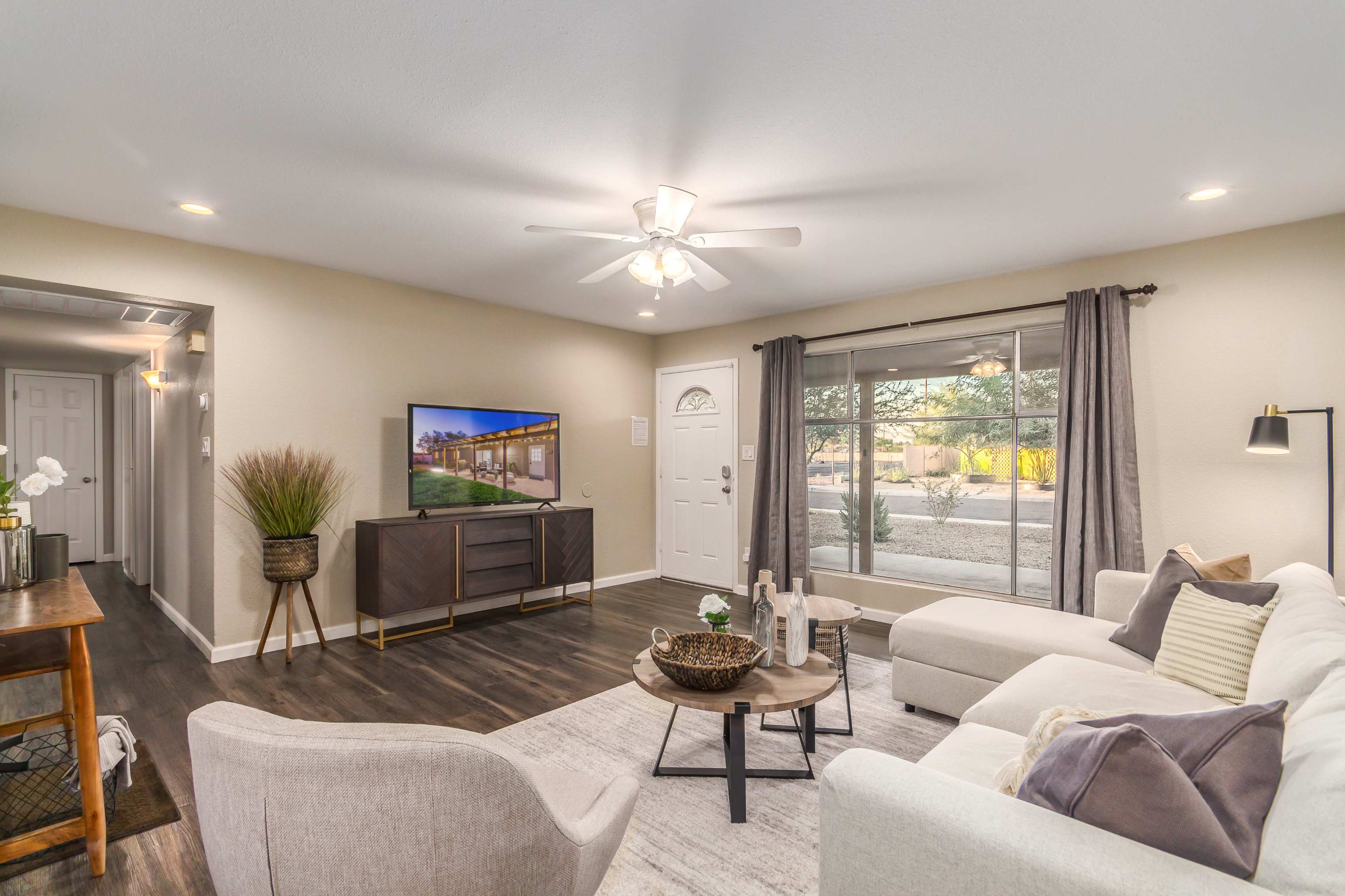 A modern living room features a white sectional sofa, a coffee table, and a television on a dark wood cabinet, with large windows allowing natural light.