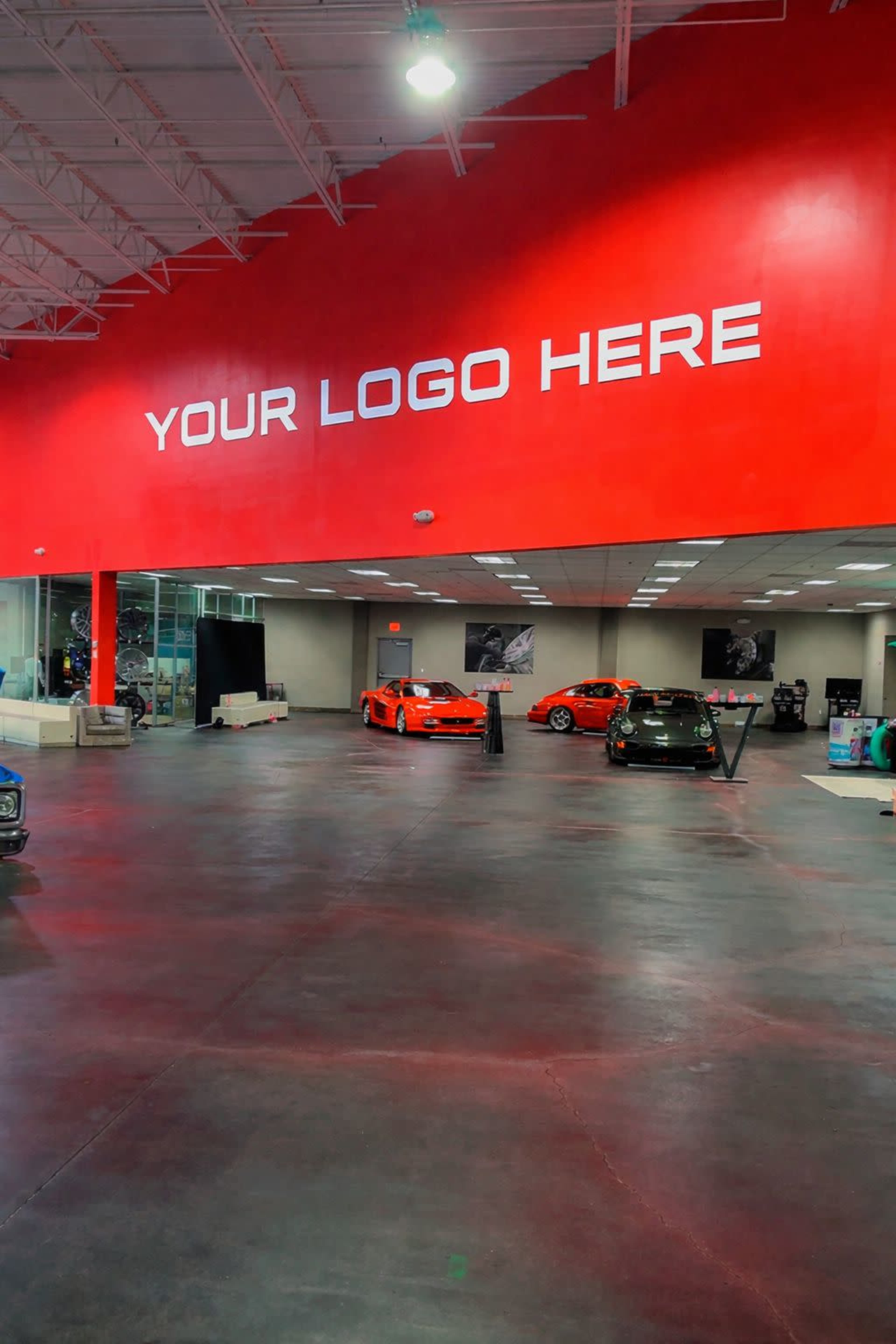 A spacious car showroom features several vehicles displayed on a polished floor, with a prominent red wall that includes placeholder text for a logo.