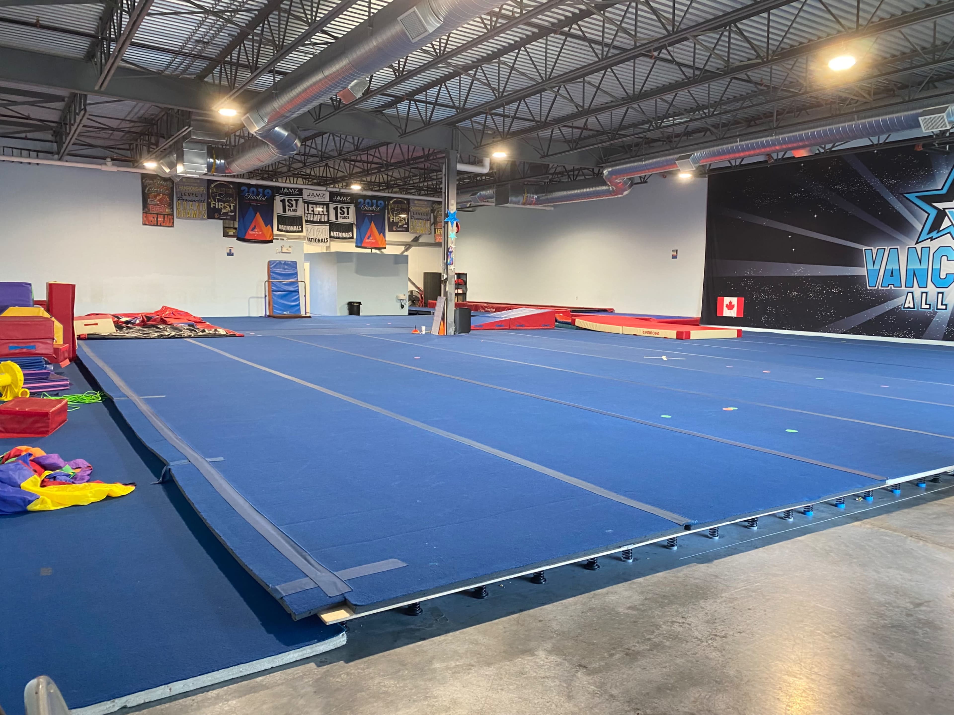 A spacious gymnasium with blue flooring, equipped for gymnastics and cheerleading, featuring various mats and equipment along the walls.