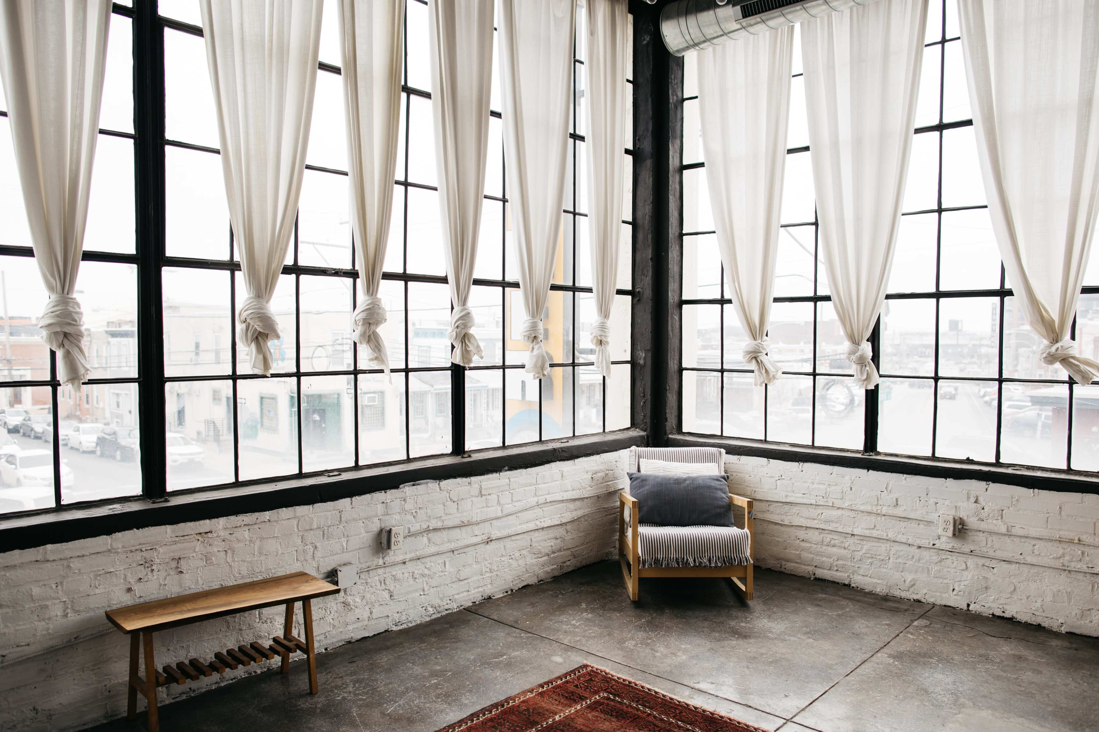 The image shows a spacious interior with large windows draped in sheer curtains, a wooden bench, and a chair facing the view outside.