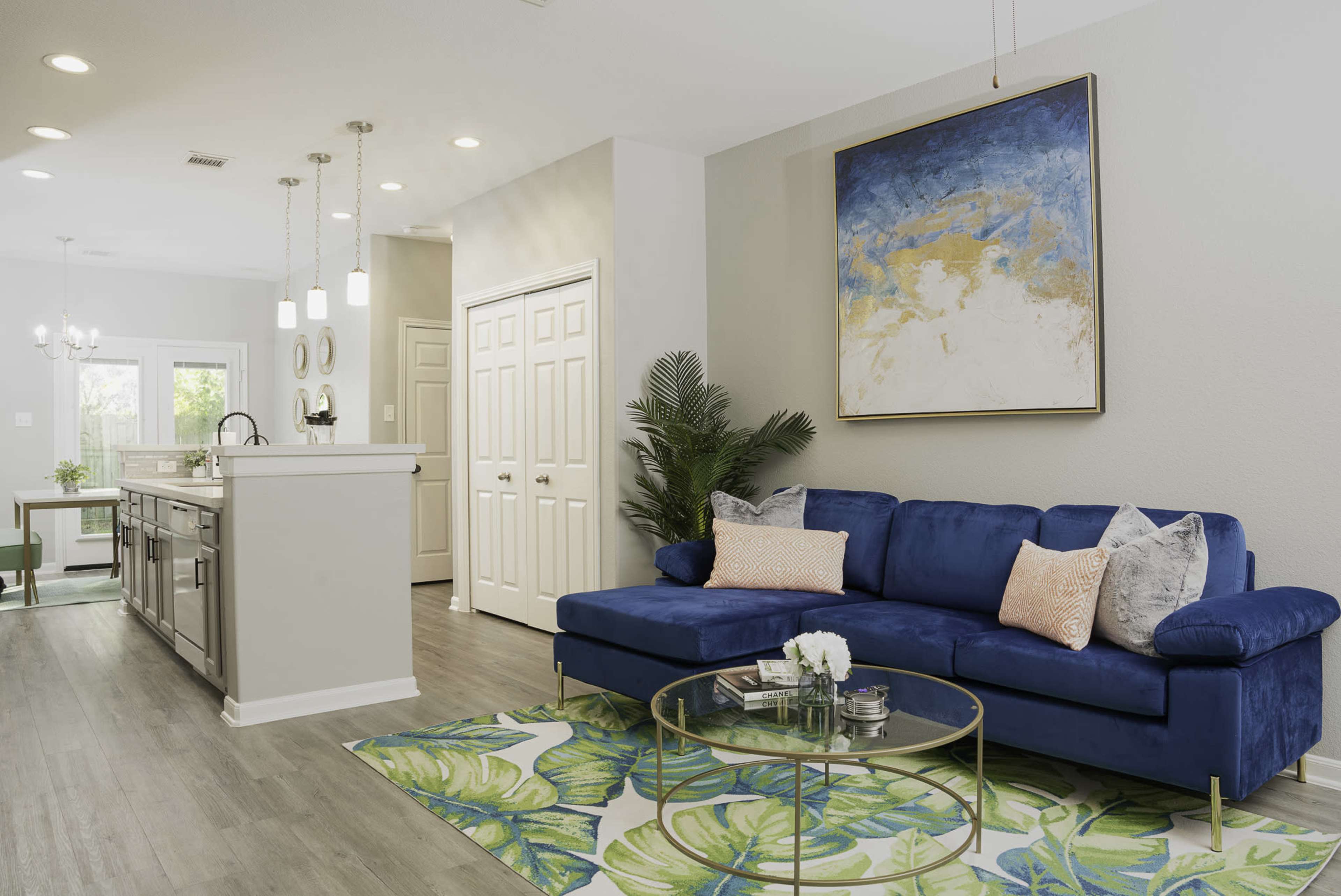 A modern living space features a blue couch, a round glass coffee table, and a large painting on the wall, with an adjacent kitchen visible in the background.