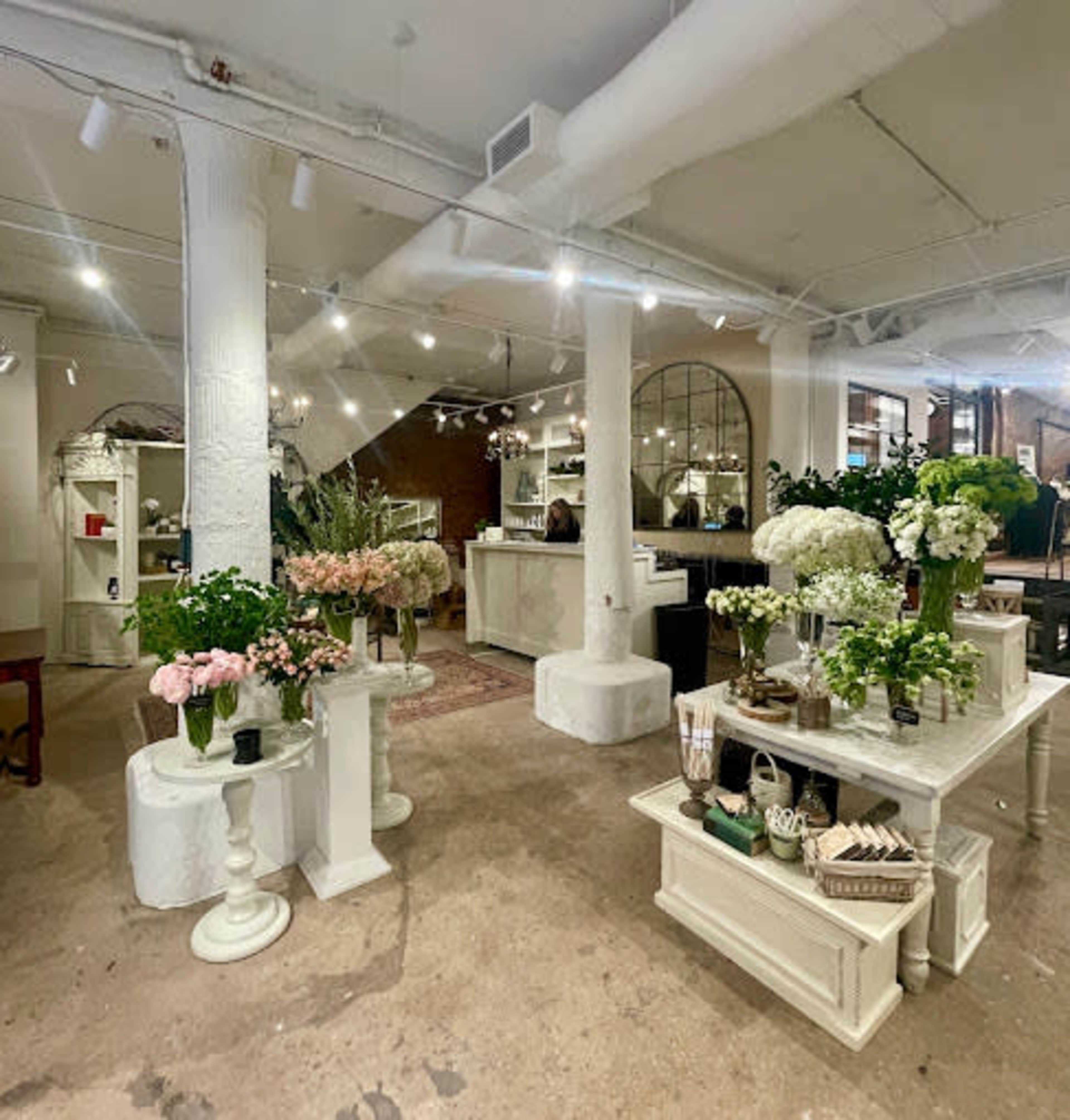 A spacious flower shop features various floral arrangements displayed on white tables within a well-lit interior.