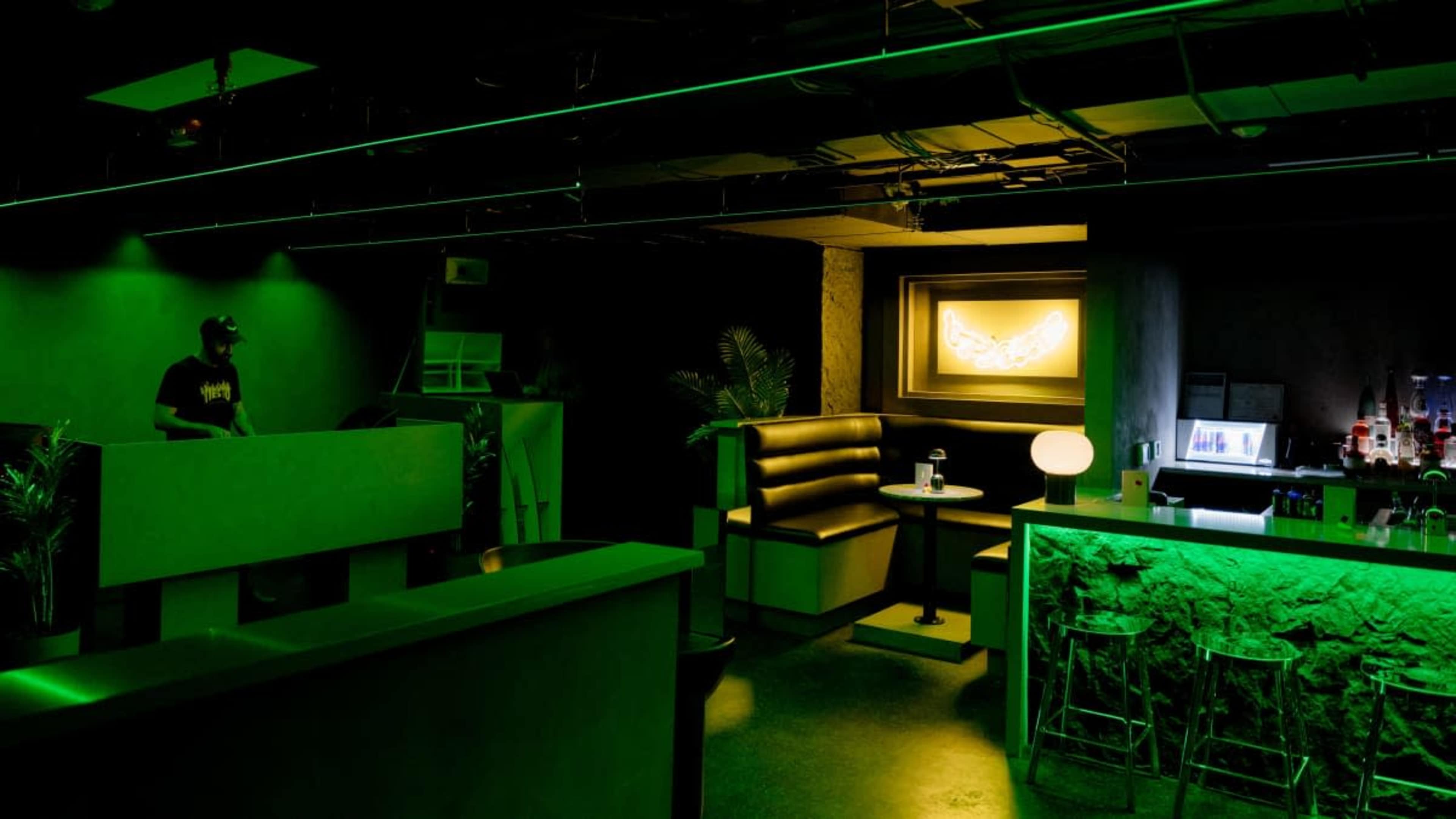 A dimly lit lounge features green lighting, minimalist furniture, and a bar area with illuminated decor.