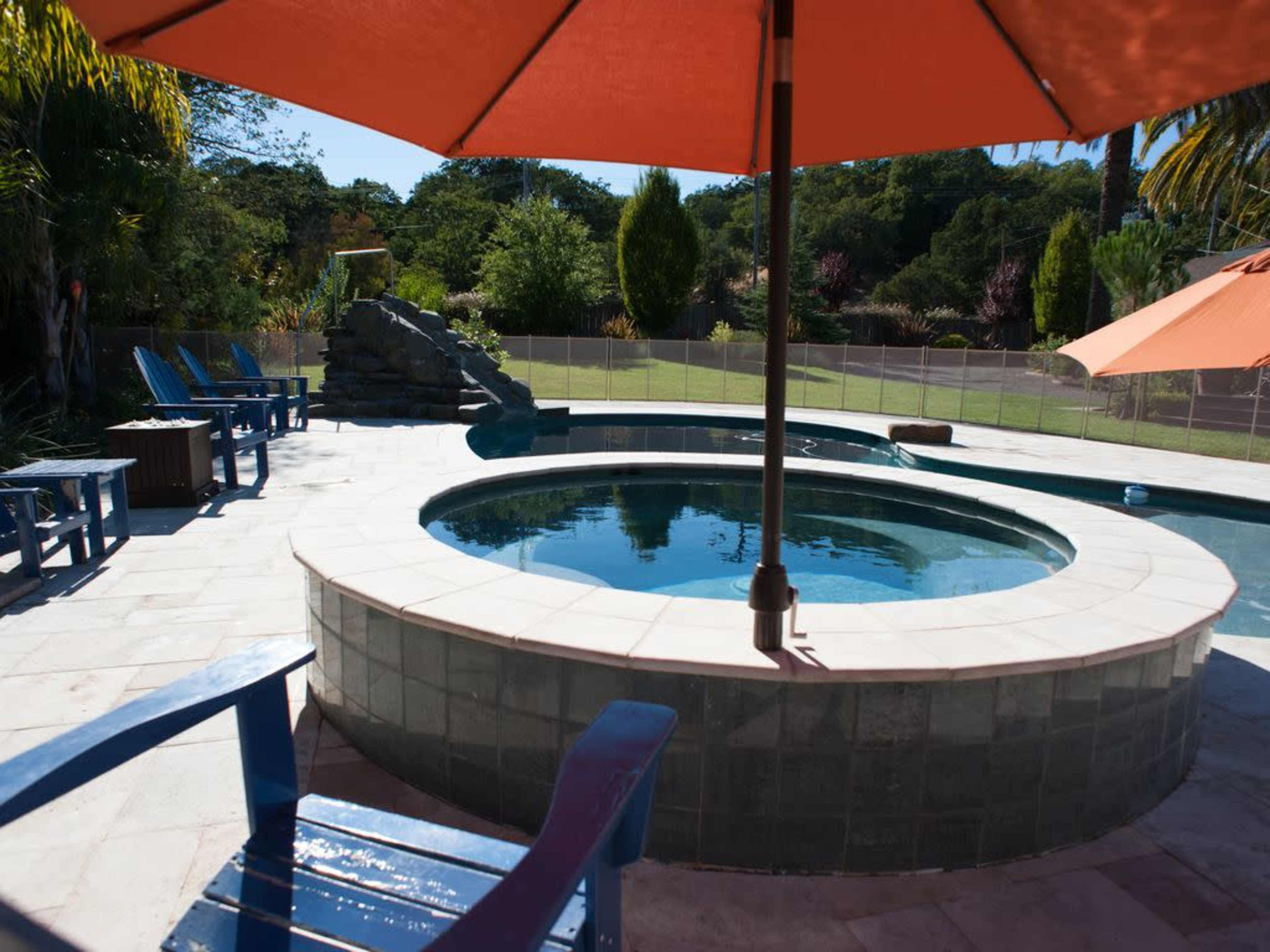 The image shows a circular hot tub surrounded by blue Adirondack chairs and orange umbrellas in a landscaped backyard.