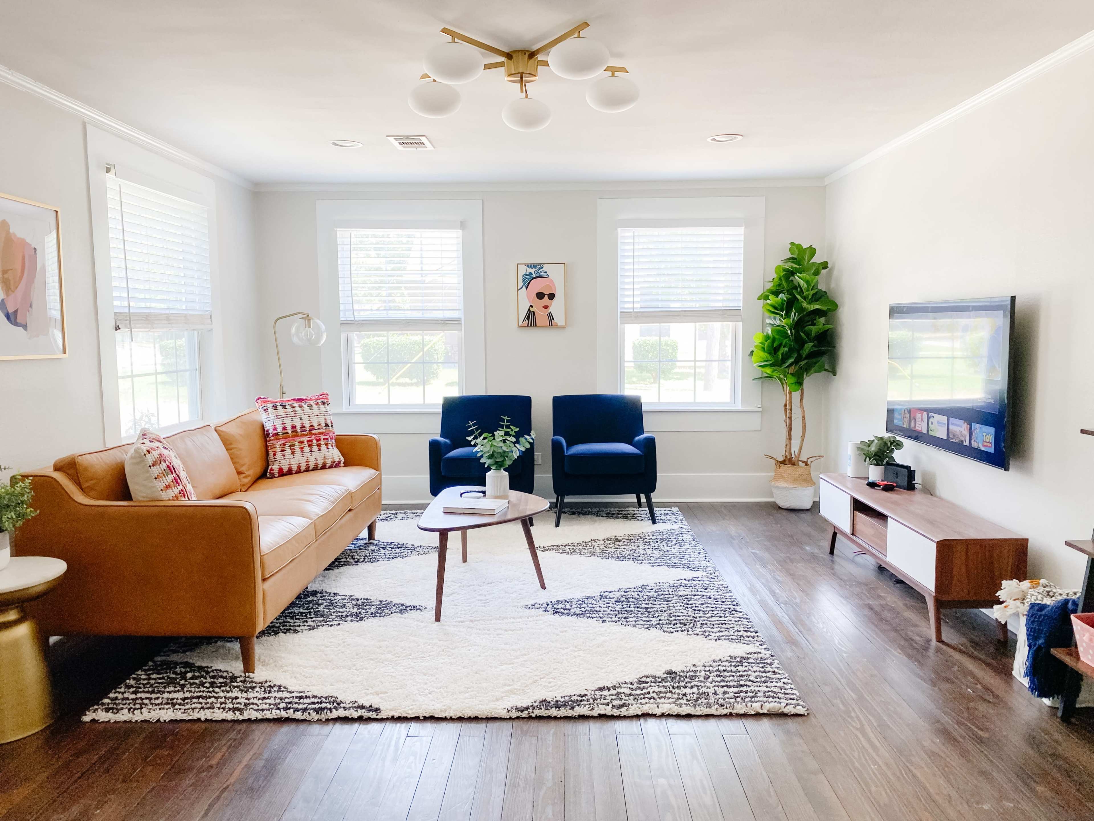 The living room features a tan leather sofa, two blue armchairs, a coffee table, and a patterned rug, illuminated by a modern ceiling light.