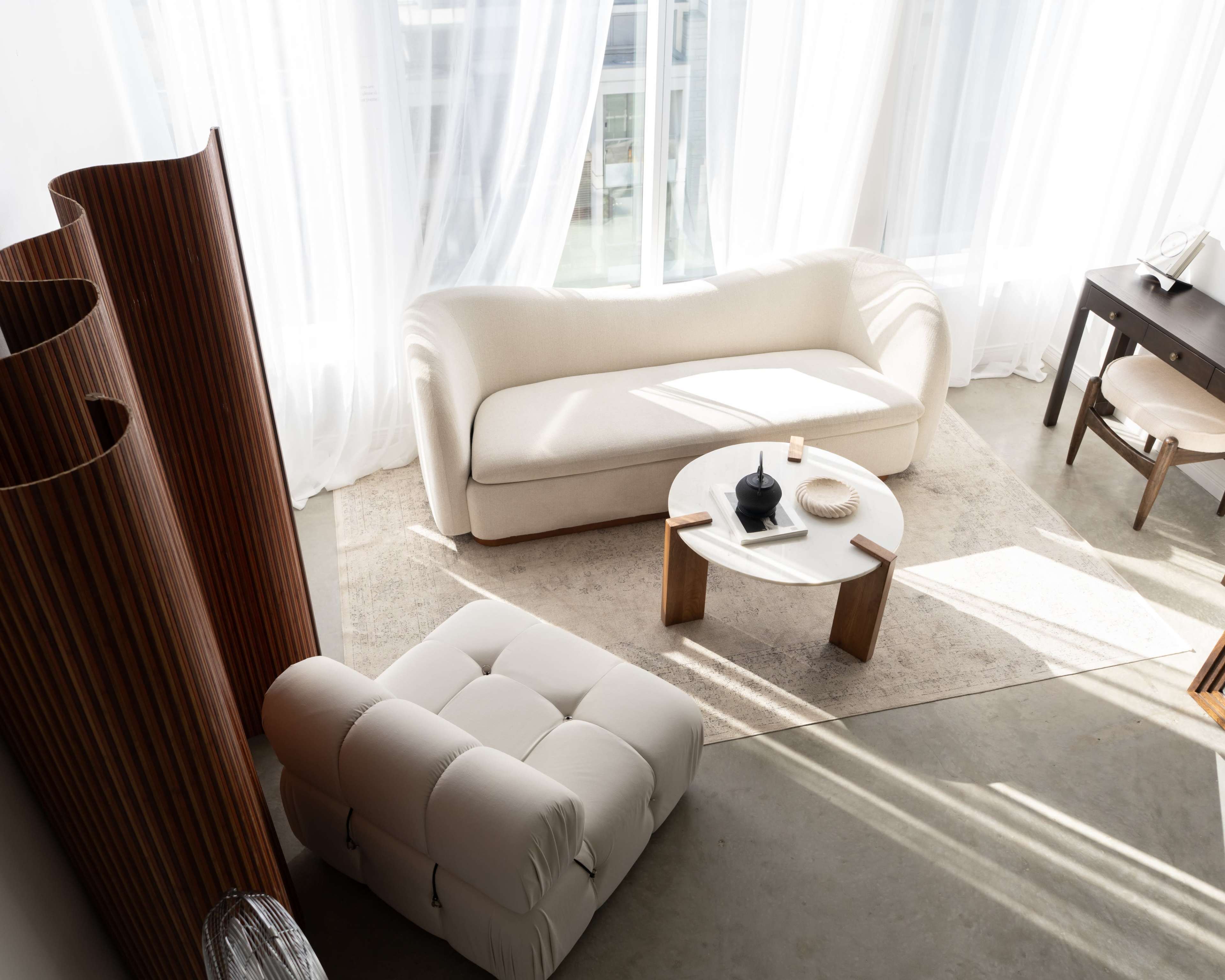 The image depicts a modern living room featuring a cream-colored sofa, a round coffee table, and a light-colored rug, with large windows allowing natural light to flood the space.