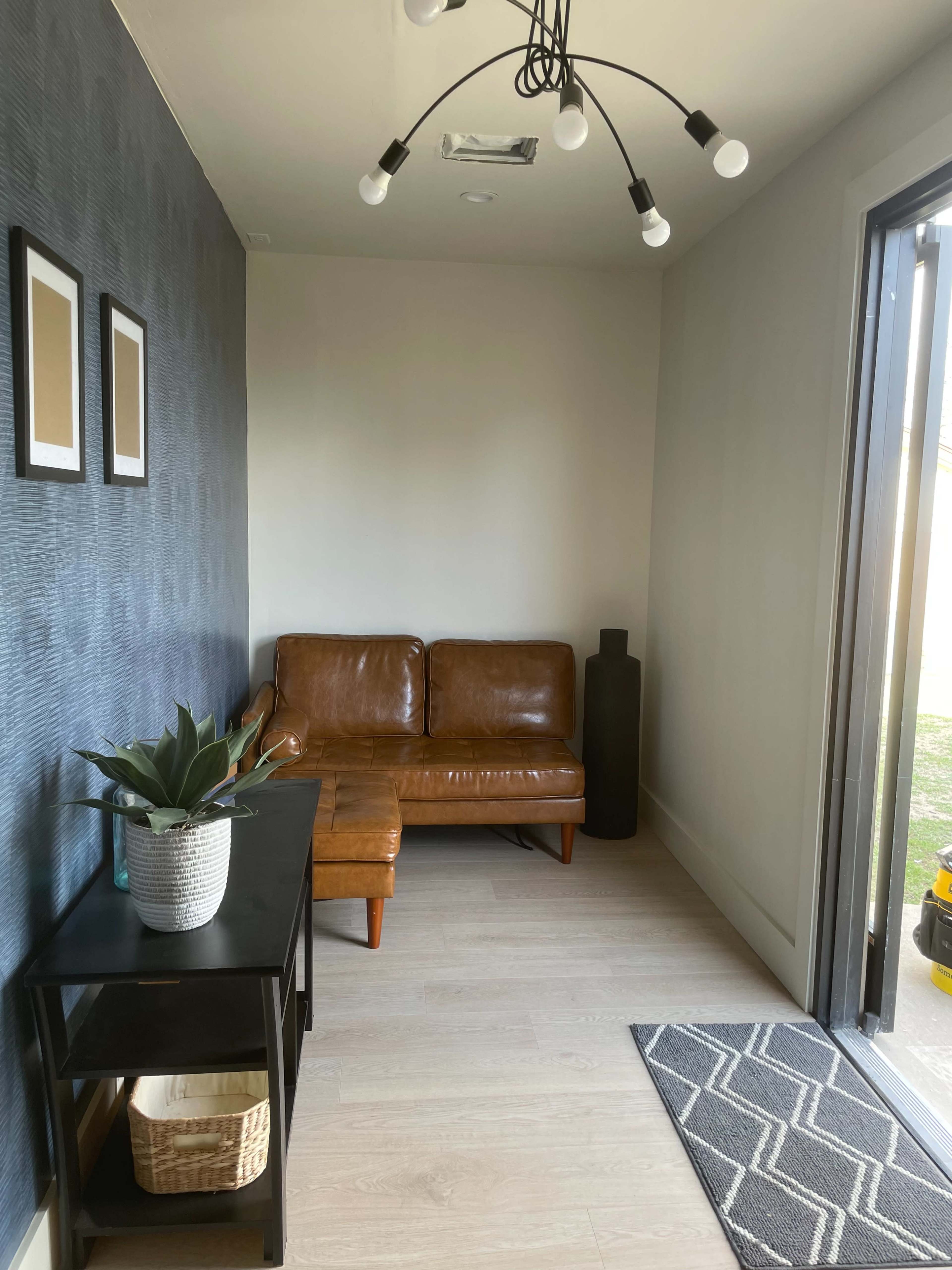 The image shows a small alcove with a brown leather sofa, a black side table, a potted plant, and two framed pictures on the wall, with a door leading outside.