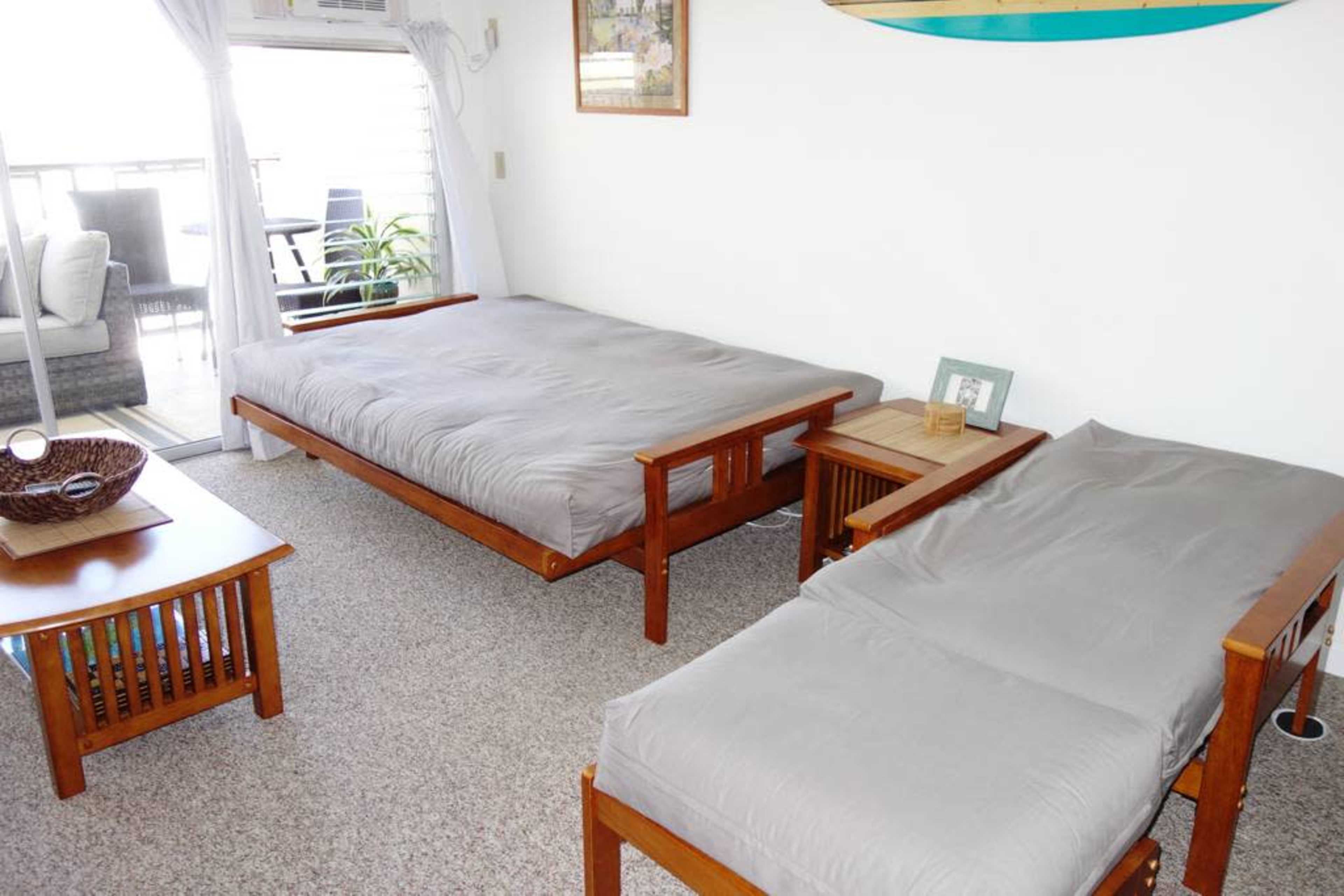 The room features two wooden futon frames with gray mattresses and a small wooden table between them, set against a light-colored wall and carpeted floor.