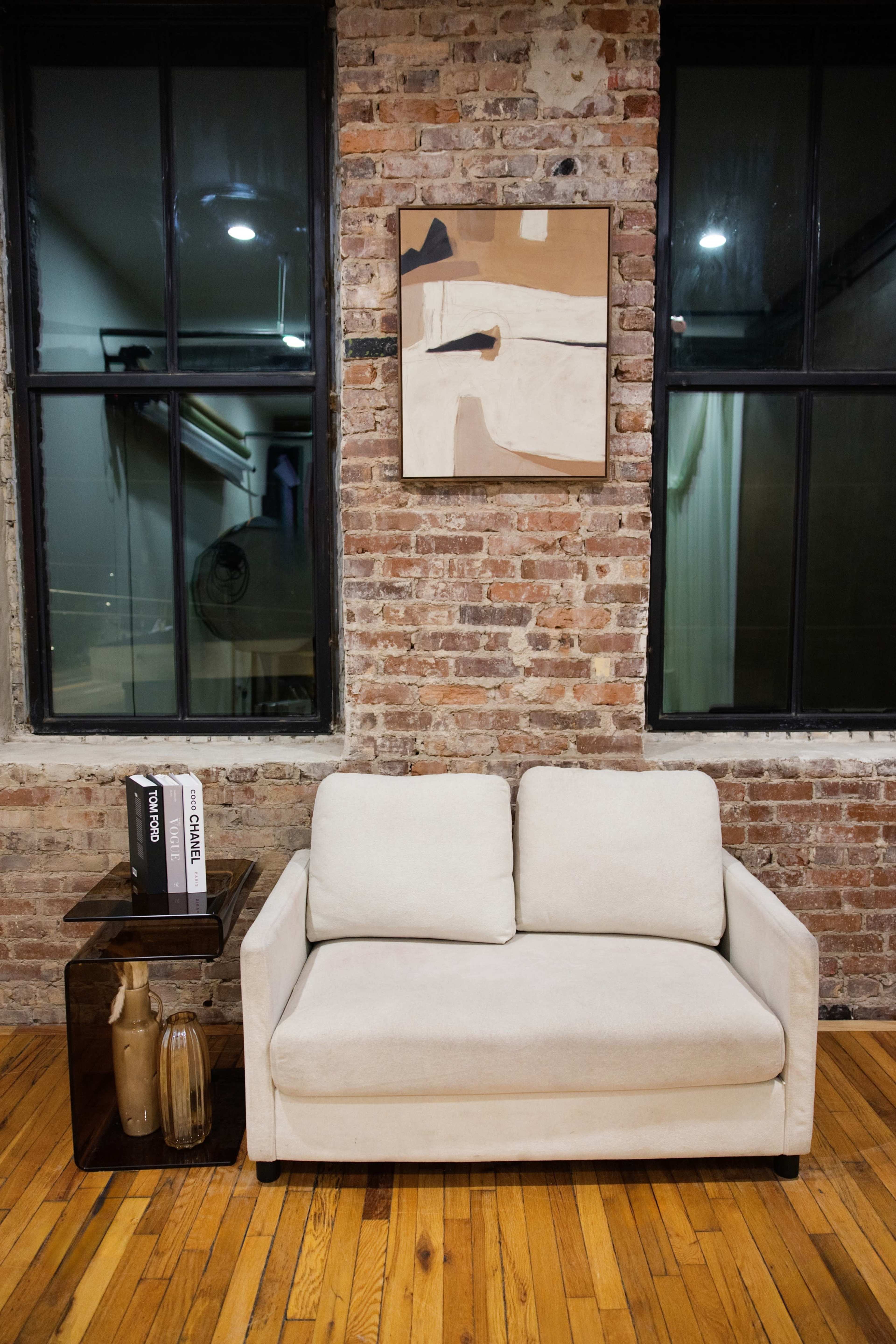 A light-colored sofa is positioned against a brick wall, next to a dark side table with a decorative vase and books, below a painting hanging on the wall.