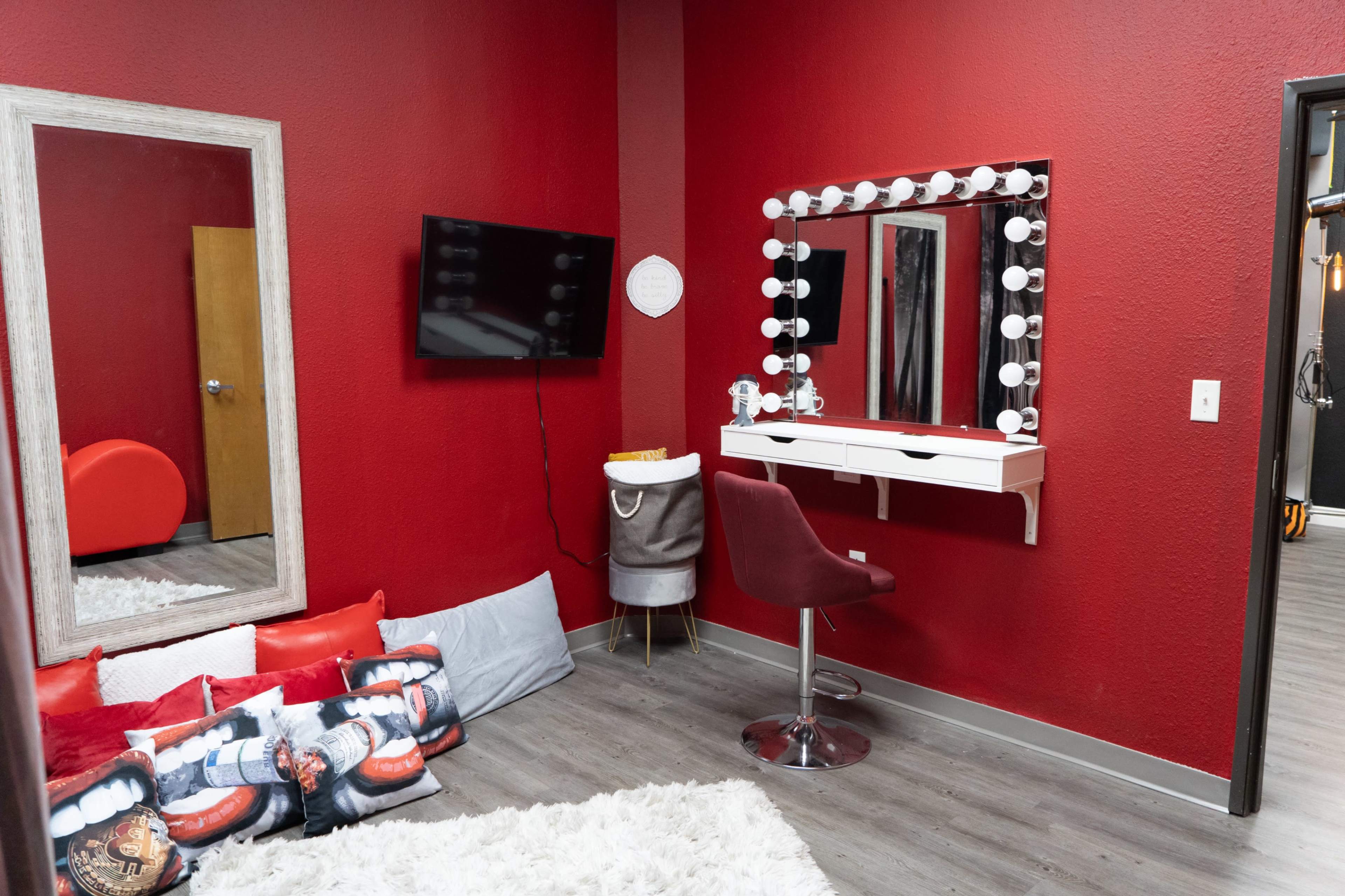 A red-walled room featuring a vanity with bright lights, a mirror, a television on the wall, and a cozy seating area with cushions on the floor.